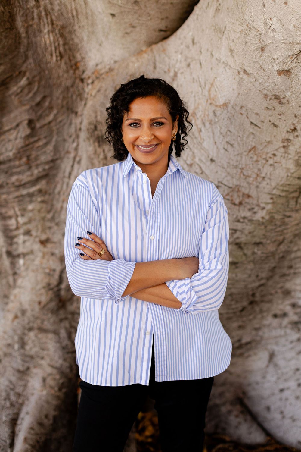 A smiling woman in a blue striped shirt stands with arms crossed before a large tree trunk.