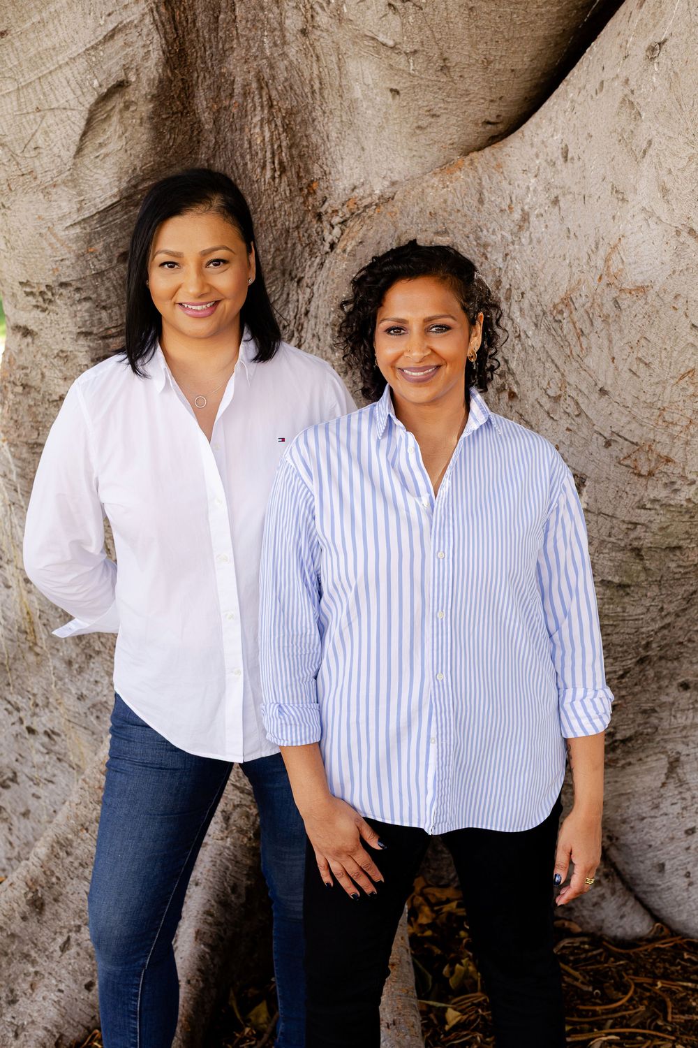 Two smiling women in button-up shirts pose together in front of a large tree trunk outdoors.