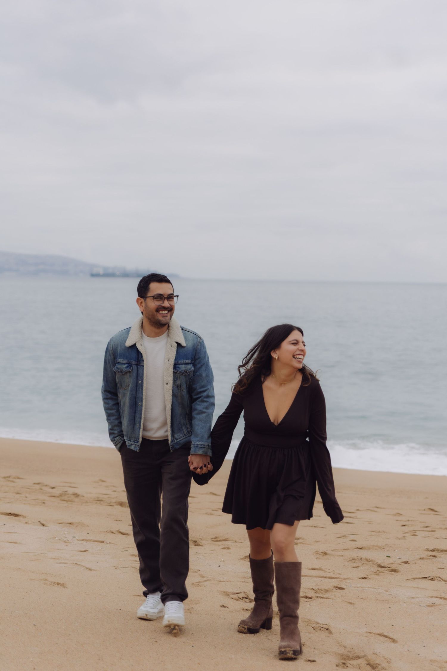 Happy couple holding hands and laughing while walking along a sandy beach on a cloudy day.