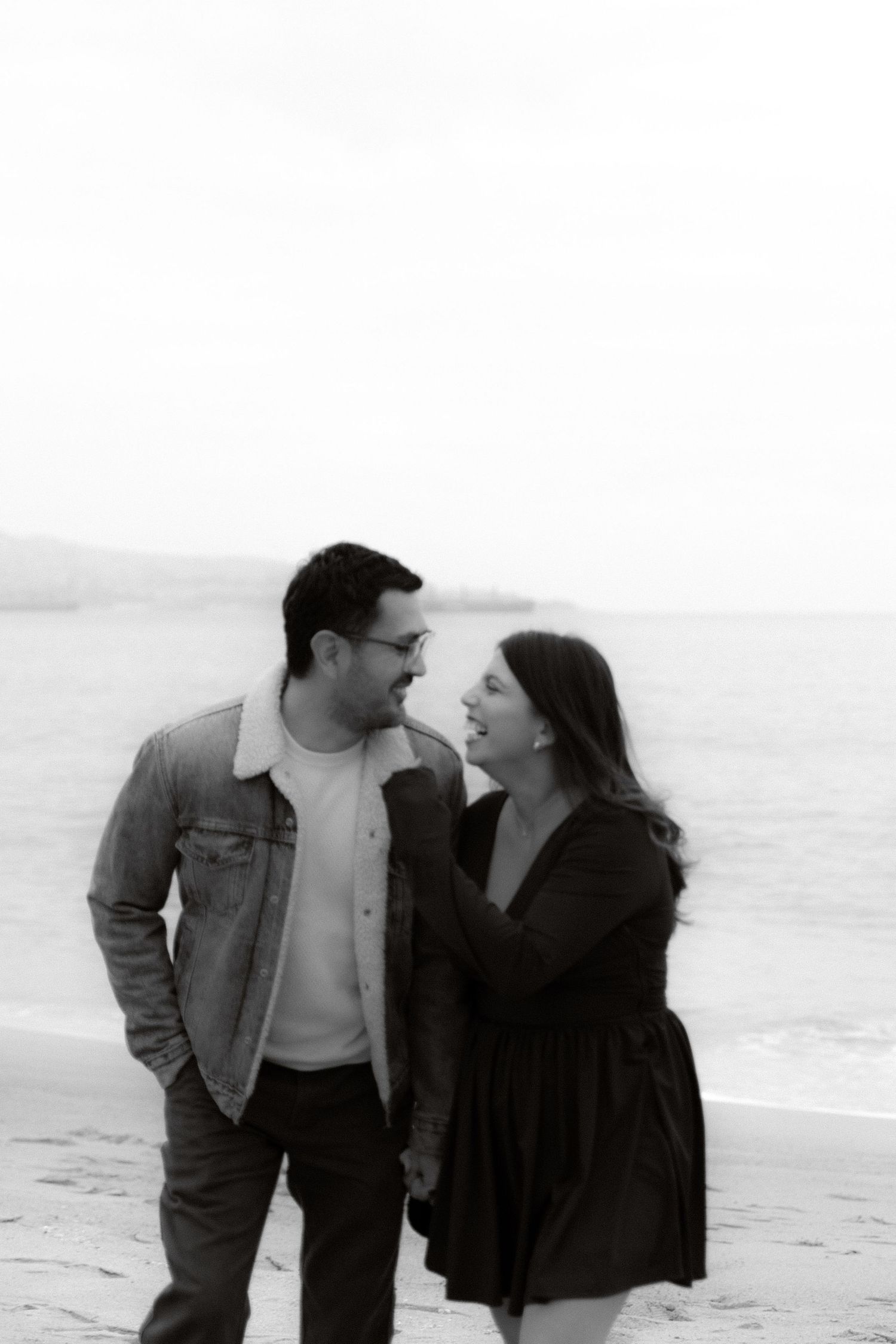 Black and white photo of a couple smiling at each other on a beach with ocean backdrop.
