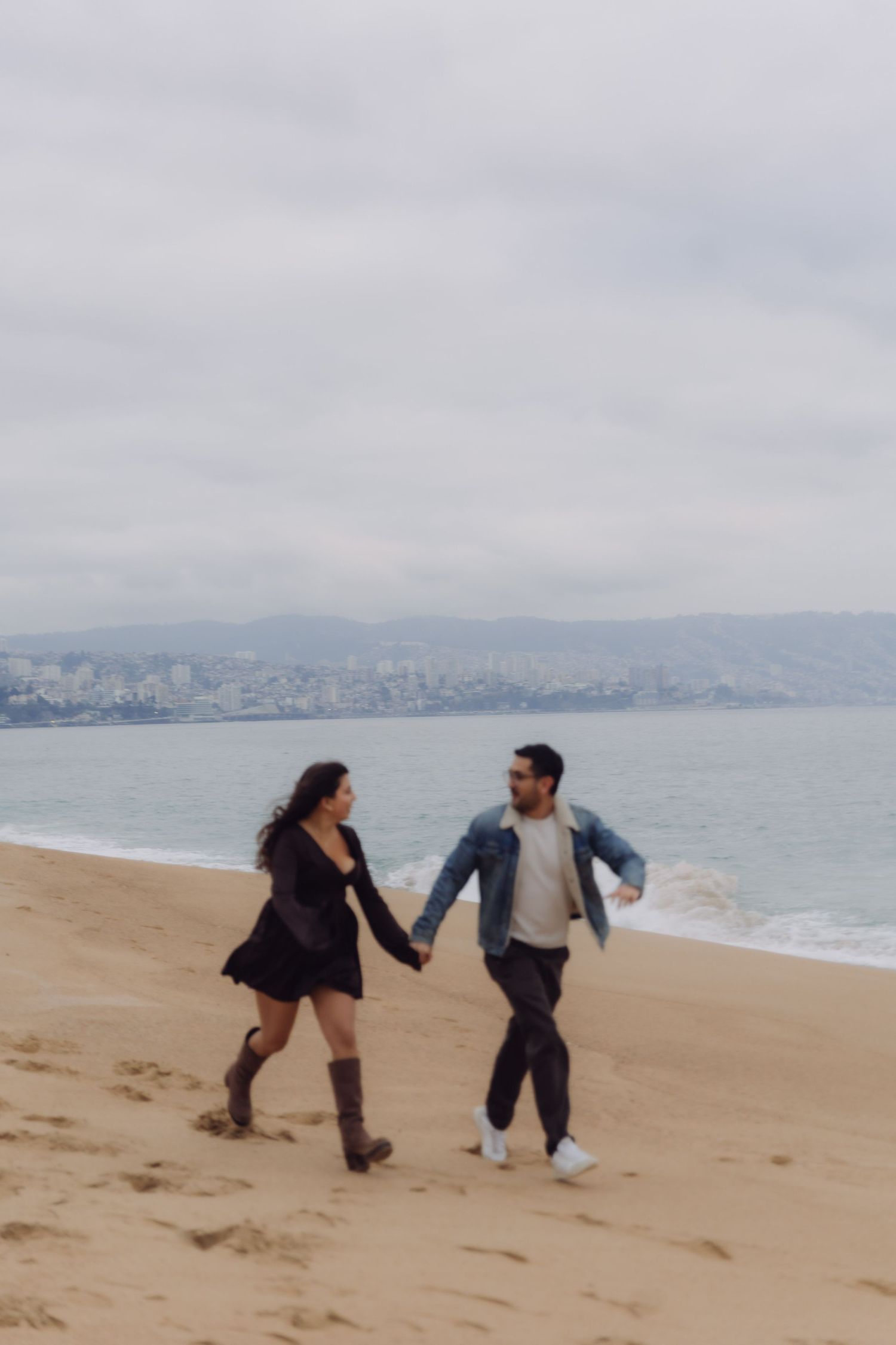 Couple holding hands and running playfully along a sandy beach with a city skyline behind them.