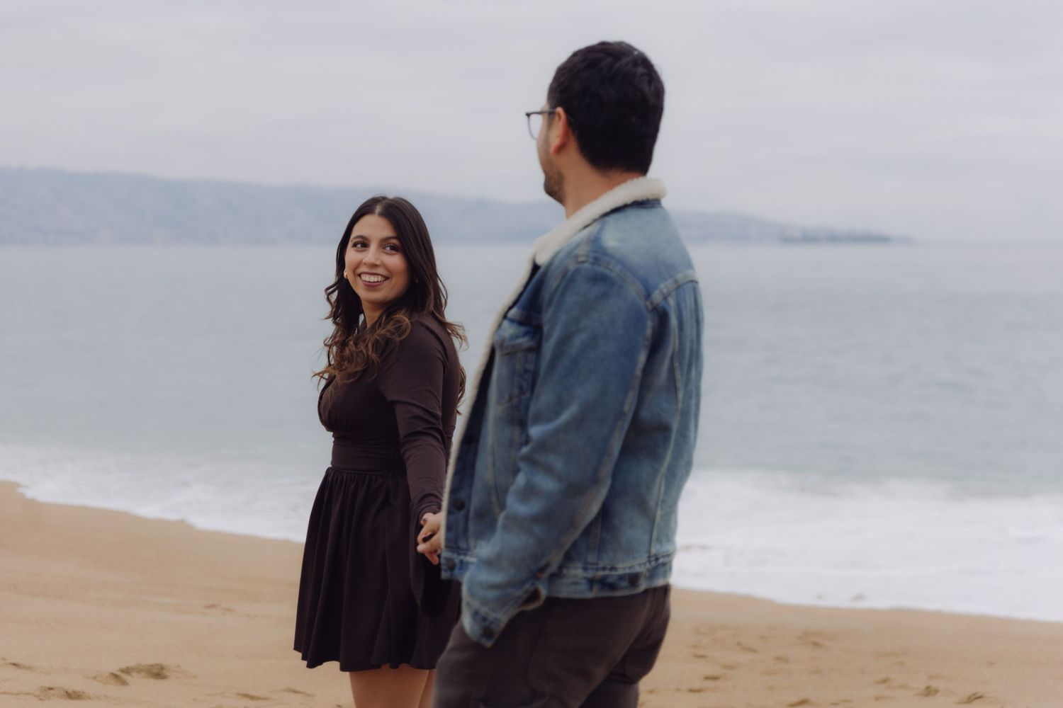 Woman in black dress glancing back at her partner while holding hands on a sandy beach.