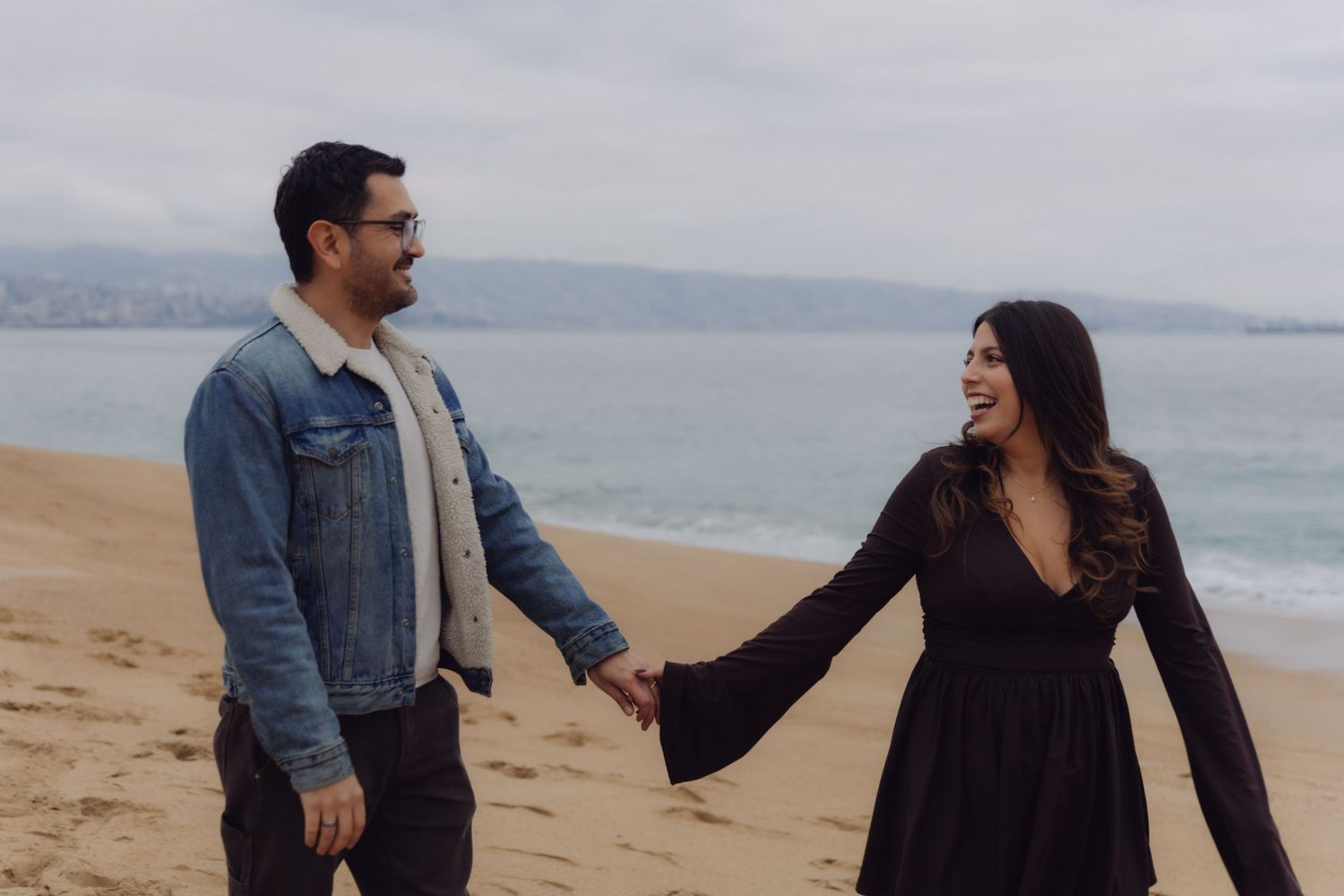Couple holding hands and smiling at each other on a sandy beach with ocean in background.