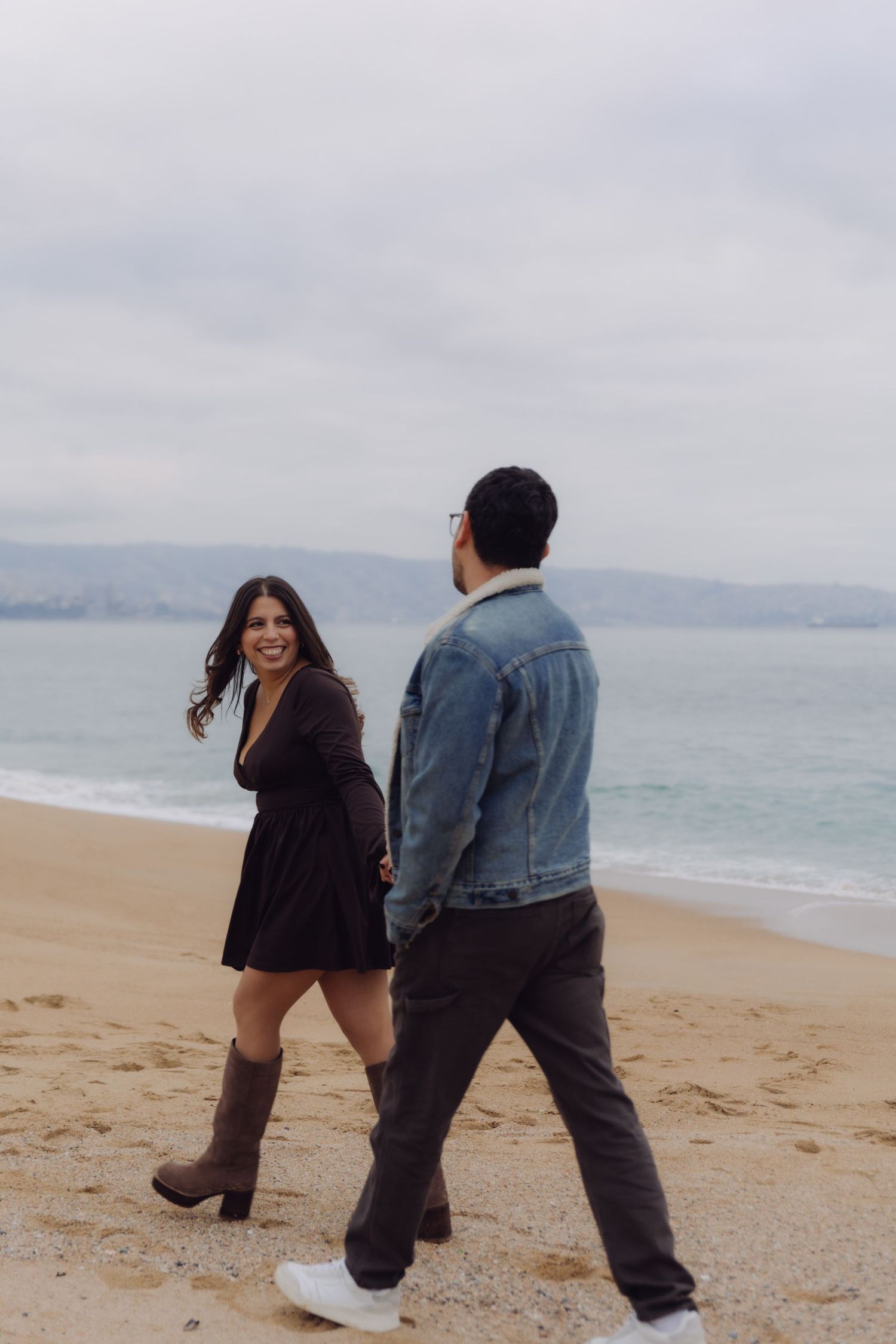 Woman in black dress and boots laughing while walking with her partner along a sandy beach.