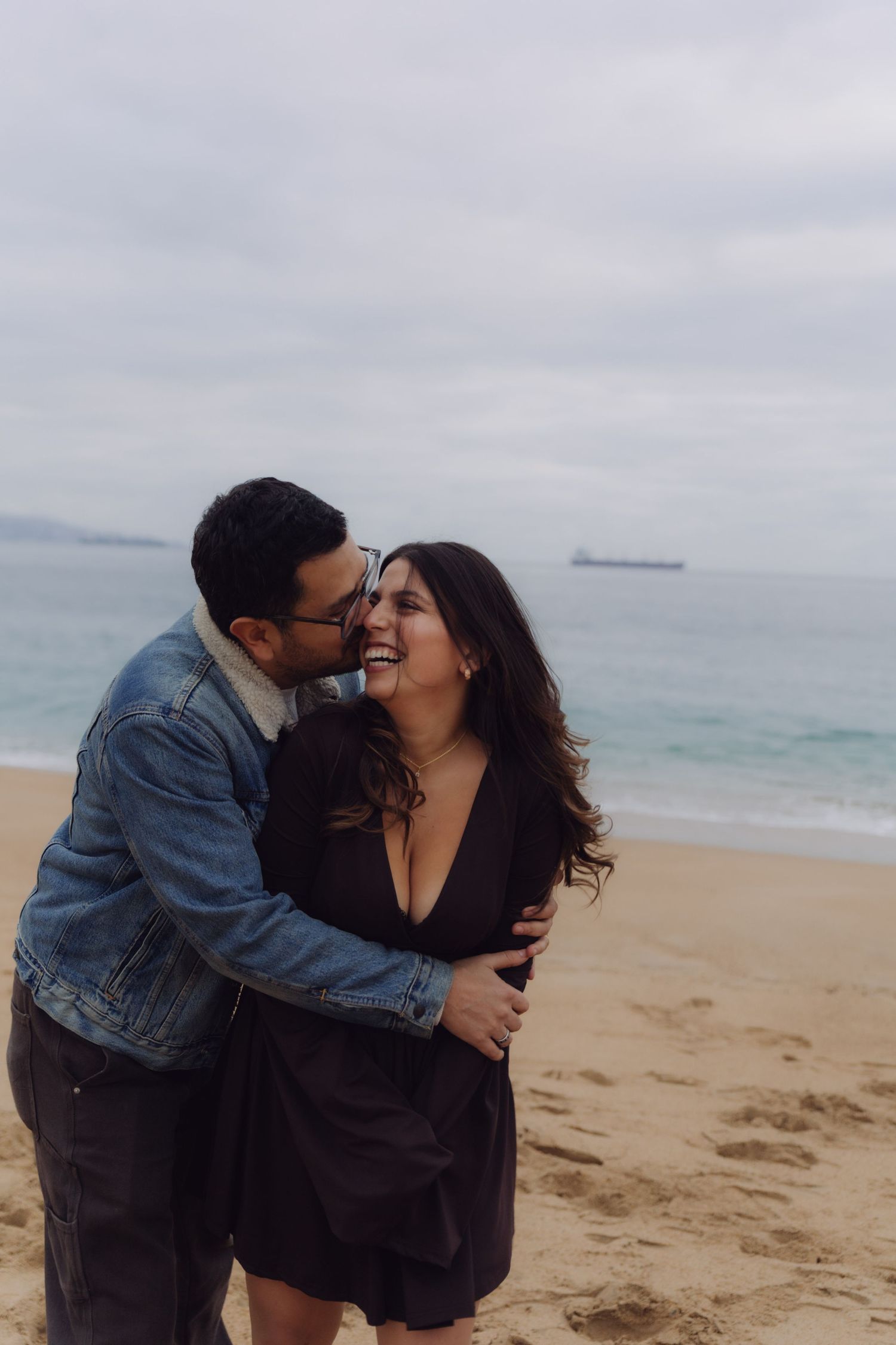 Man embracing and whispering to a laughing woman on a sandy beach with ocean behind them.