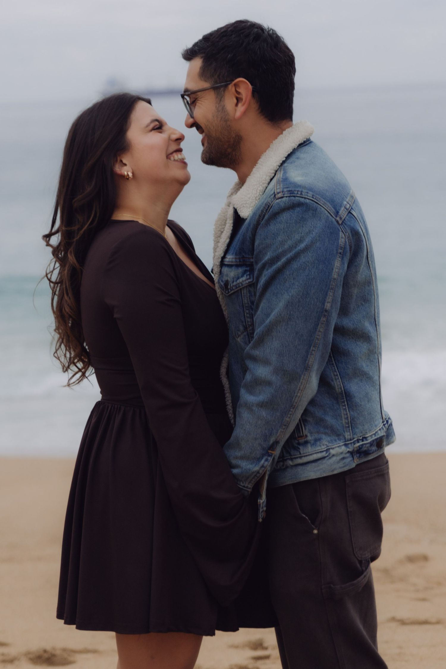 Couple smiling and gazing at each other lovingly while holding hands on a sandy beach.