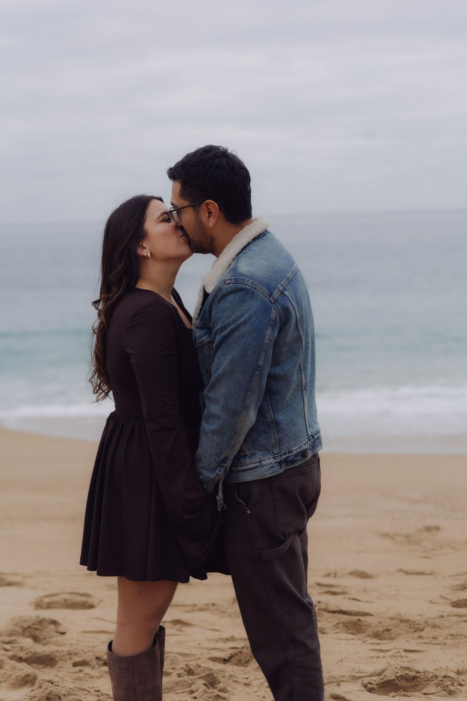Couple sharing a romantic kiss on a sandy beach with overcast skies and ocean behind them.