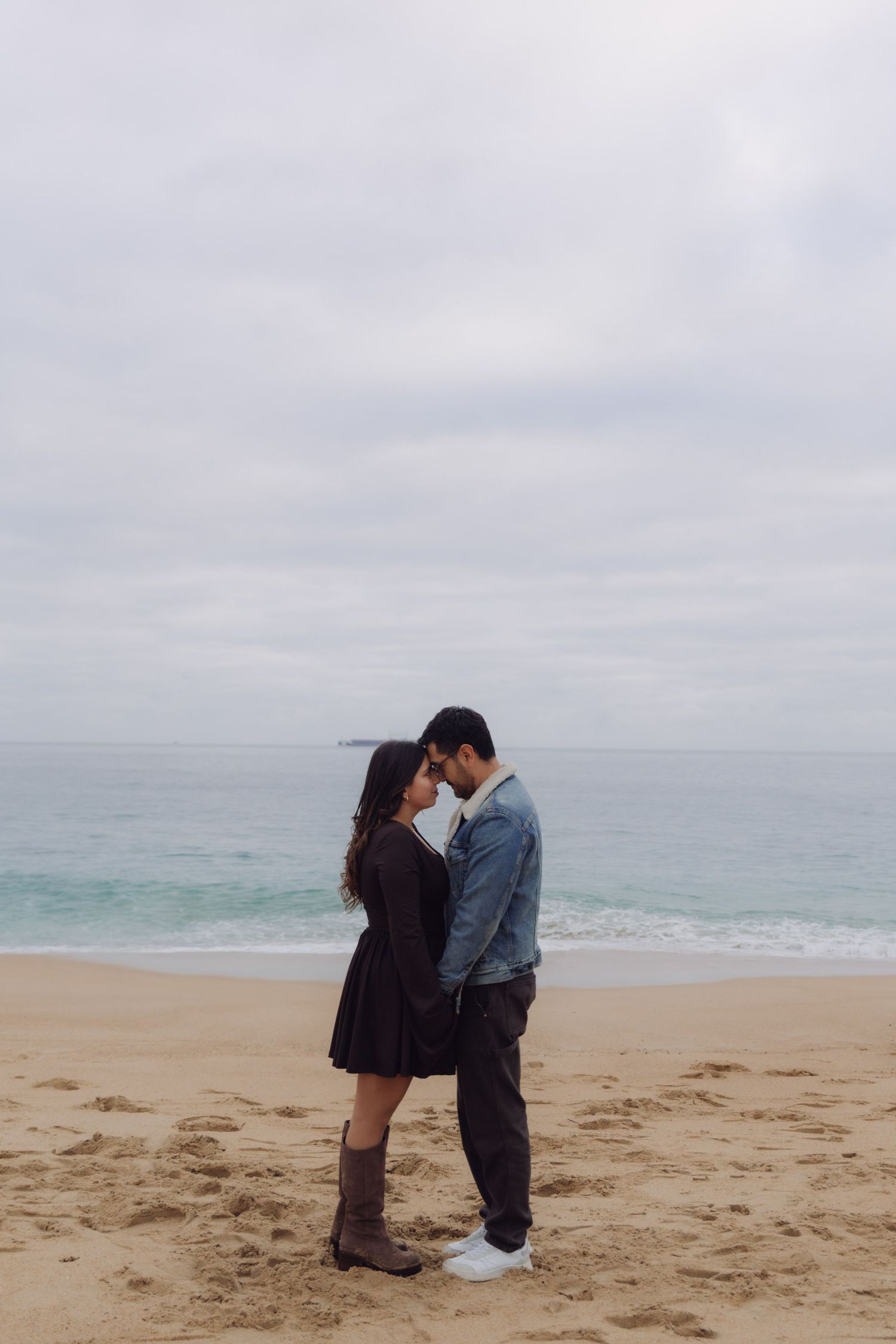 Couple embracing on a sandy beach with ocean waves in the background on a cloudy day.