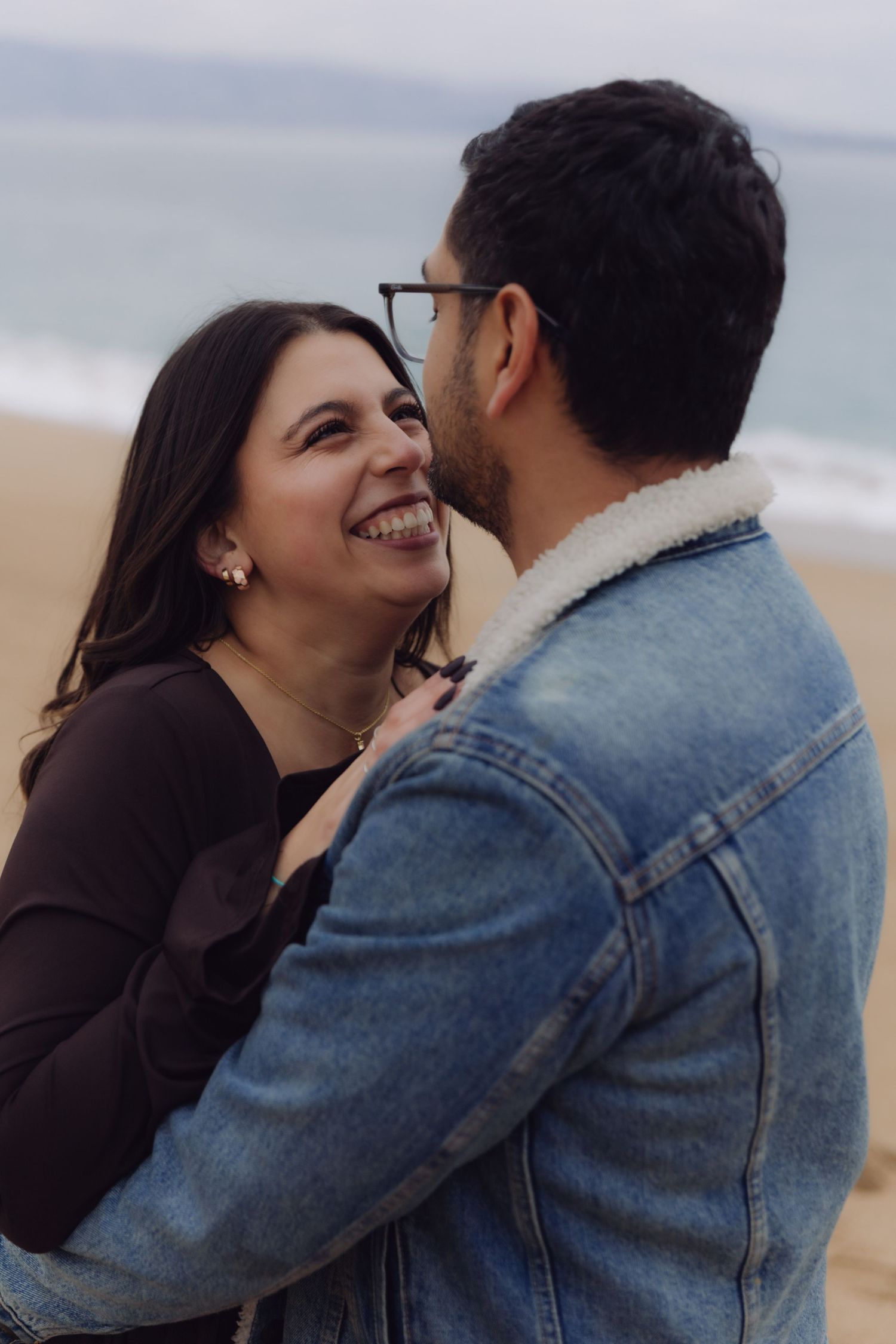 Woman laughing joyfully while embracing a man in a denim jacket on a beach.