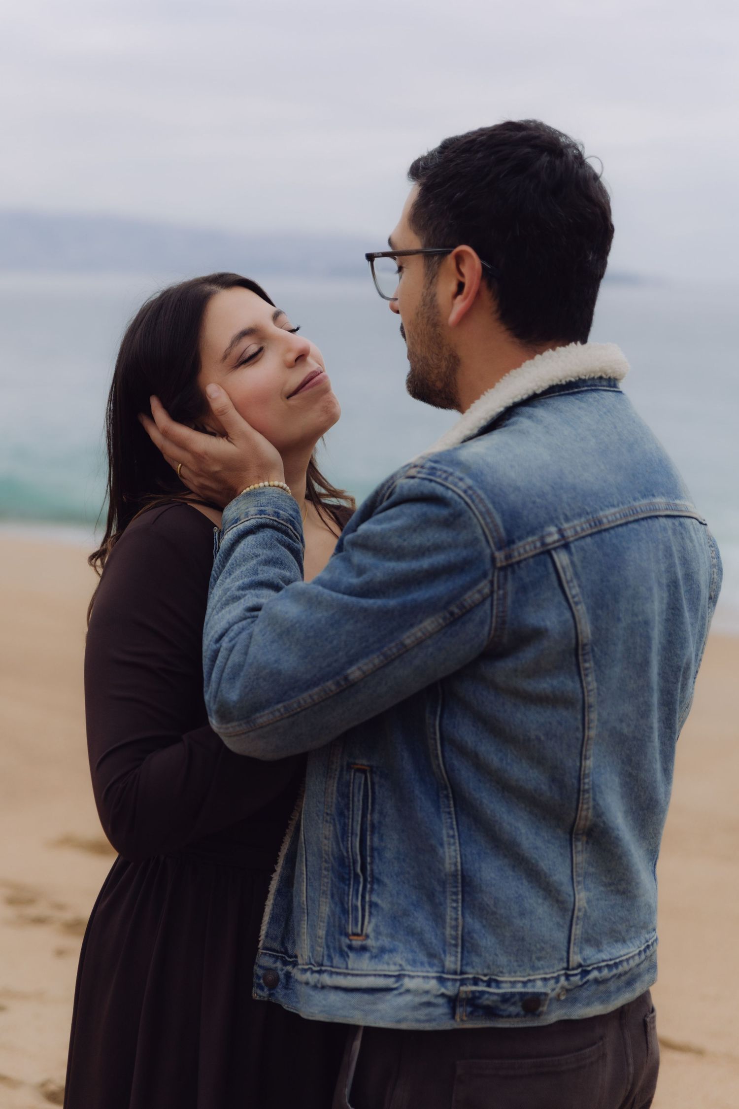 Man gently cupping womans face on a sandy beach with ocean in the background.
