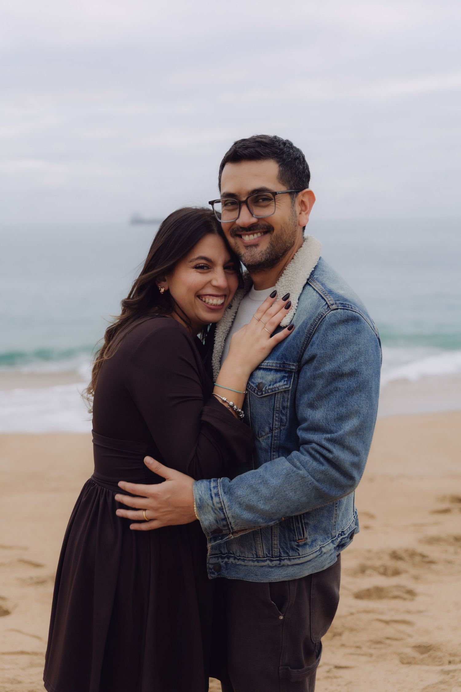 Happy couple hugging and smiling at camera on a sandy beach with ocean behind them.