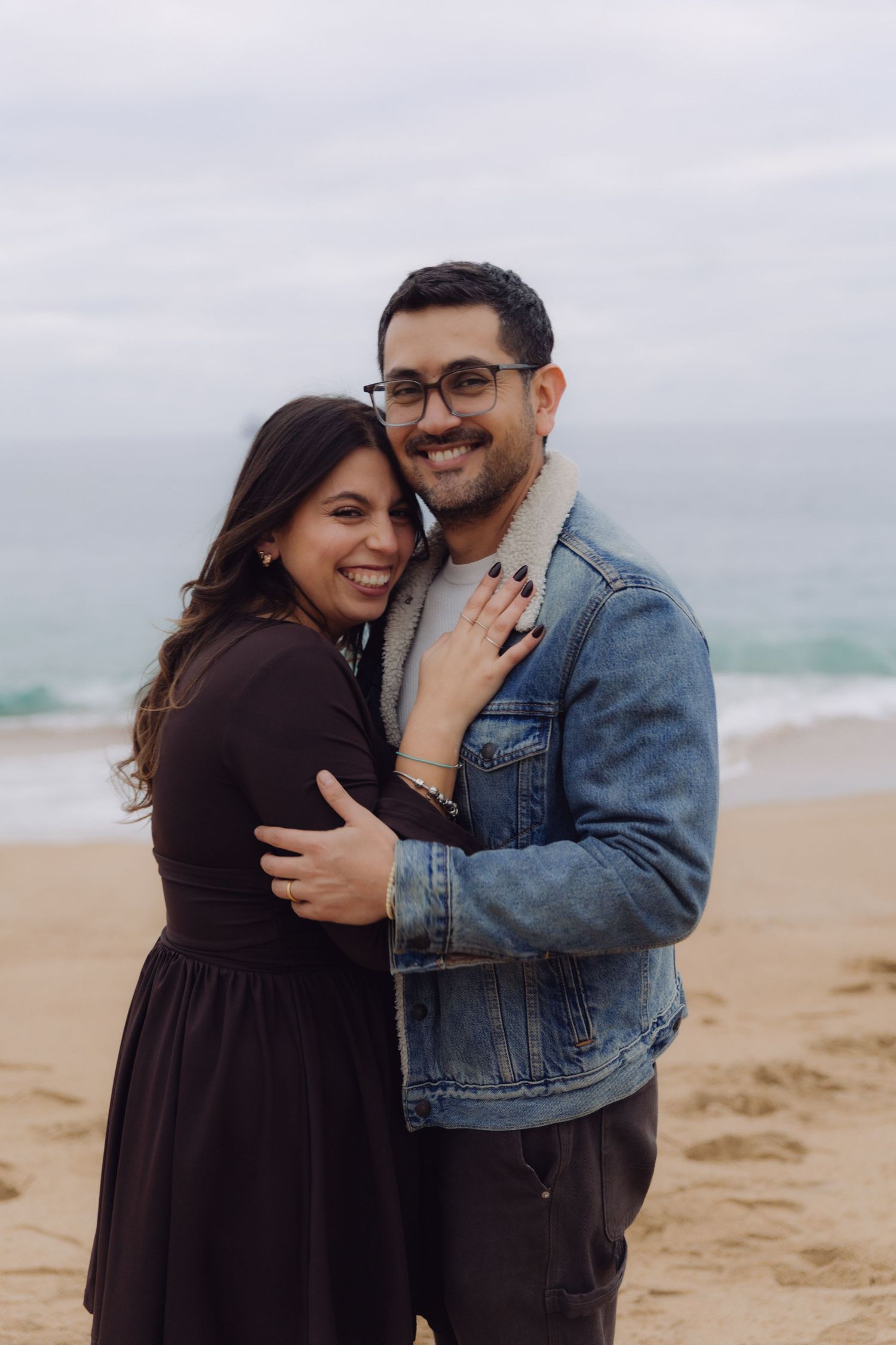 Smiling couple embracing each other on a sandy beach with waves in the background.
