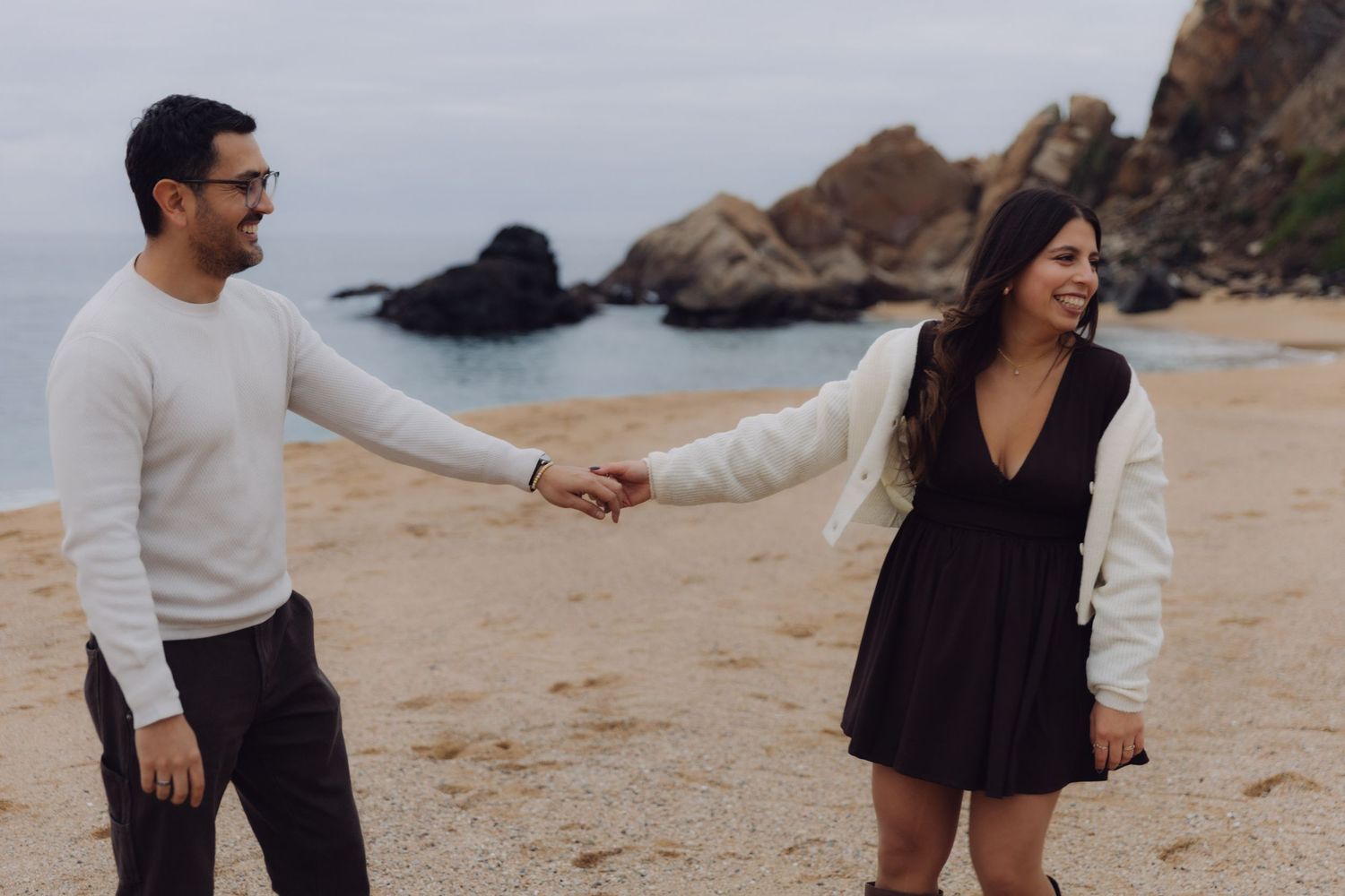 Couple holding hands and walking apart on a sandy beach with rocky cliffs behind.
