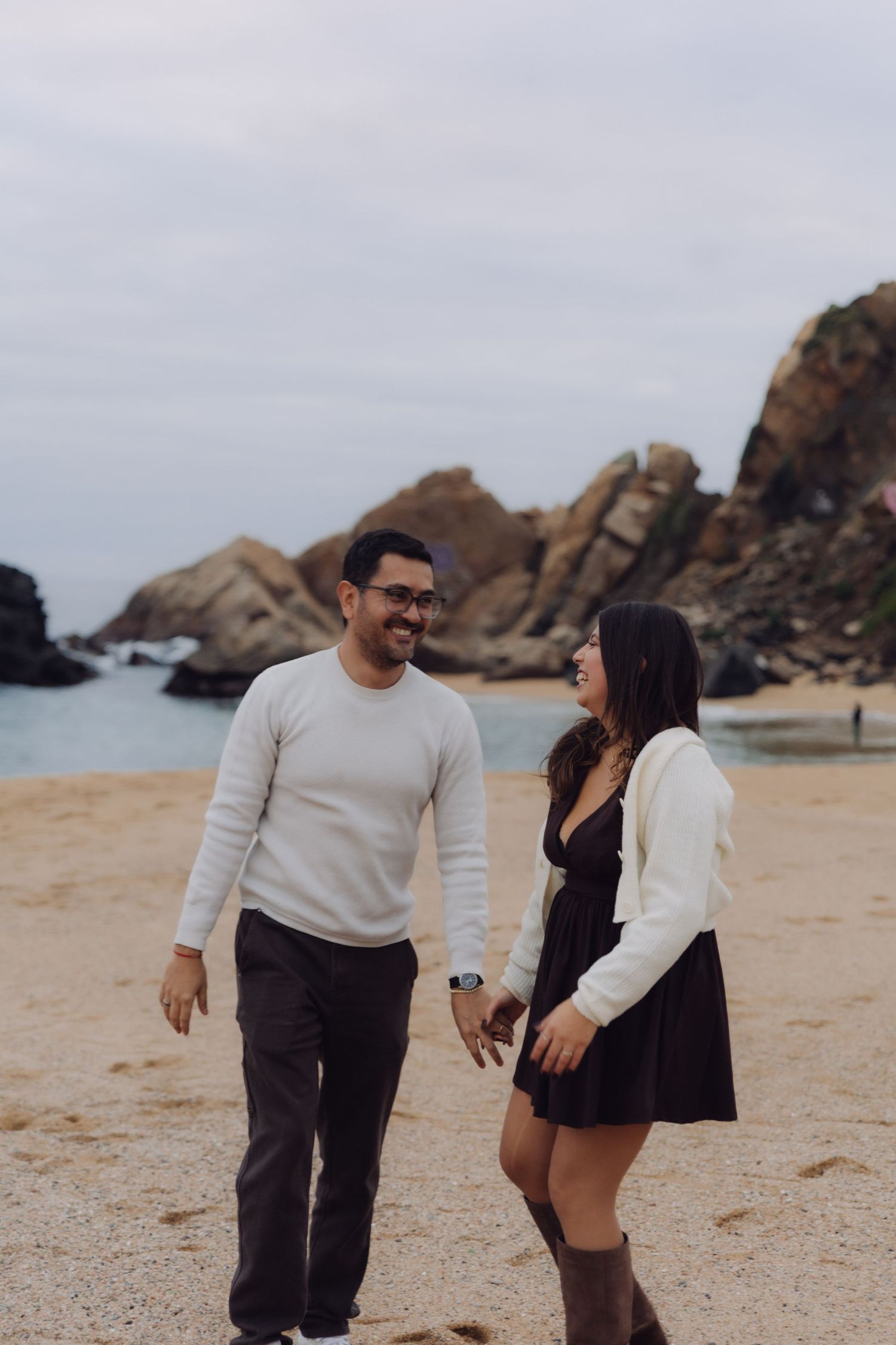 Couple walking hand in hand and laughing on a sandy beach with rocky cliffs nearby.