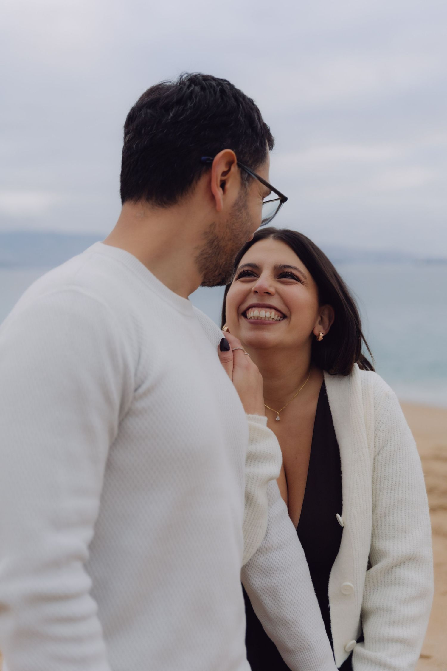 Couple laughing together on a beach, woman smiling up at man wearing glasses and white sweater.