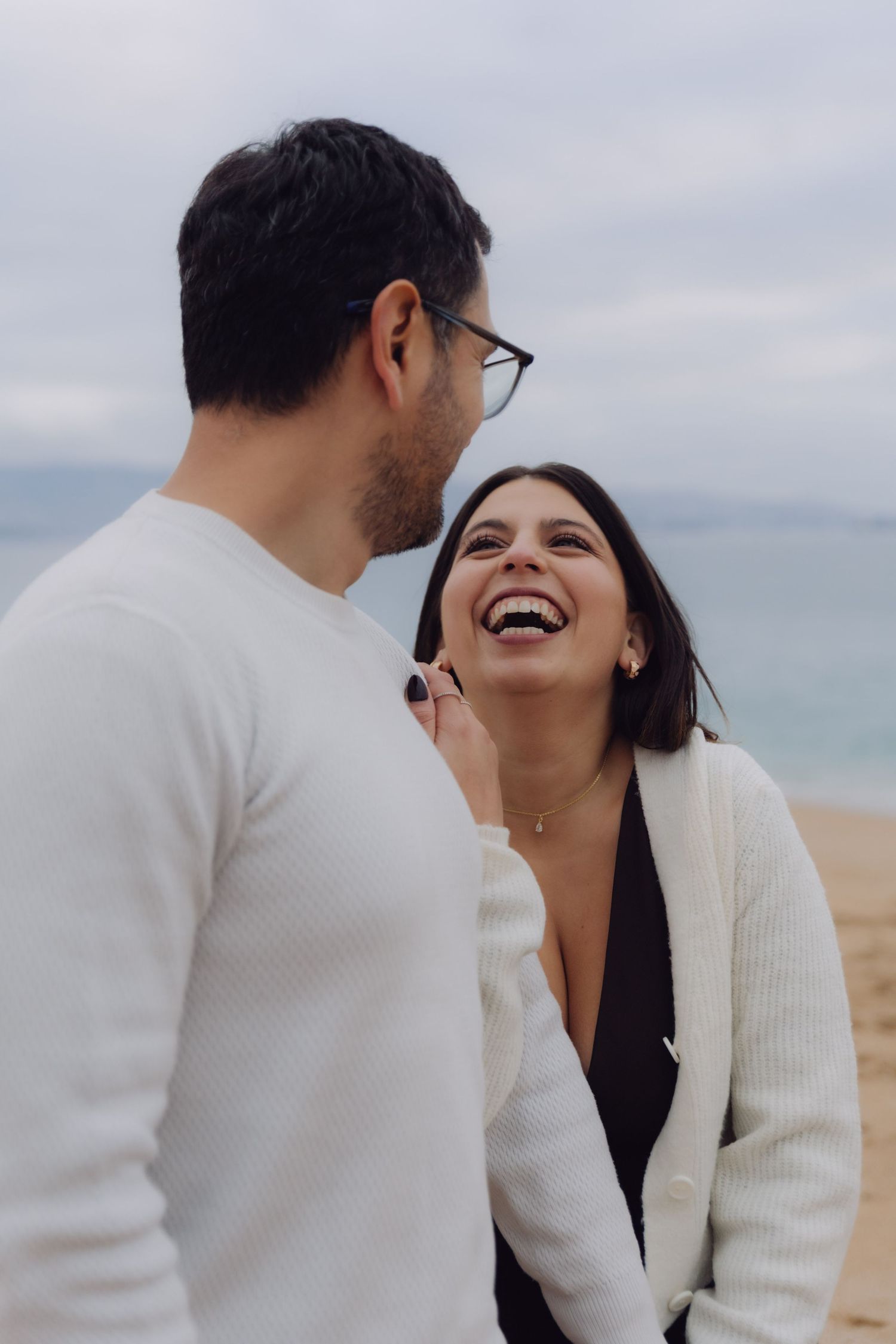 Happy couple sharing a joyful moment on a sandy beach, woman laughing in white cardigan and black dress.