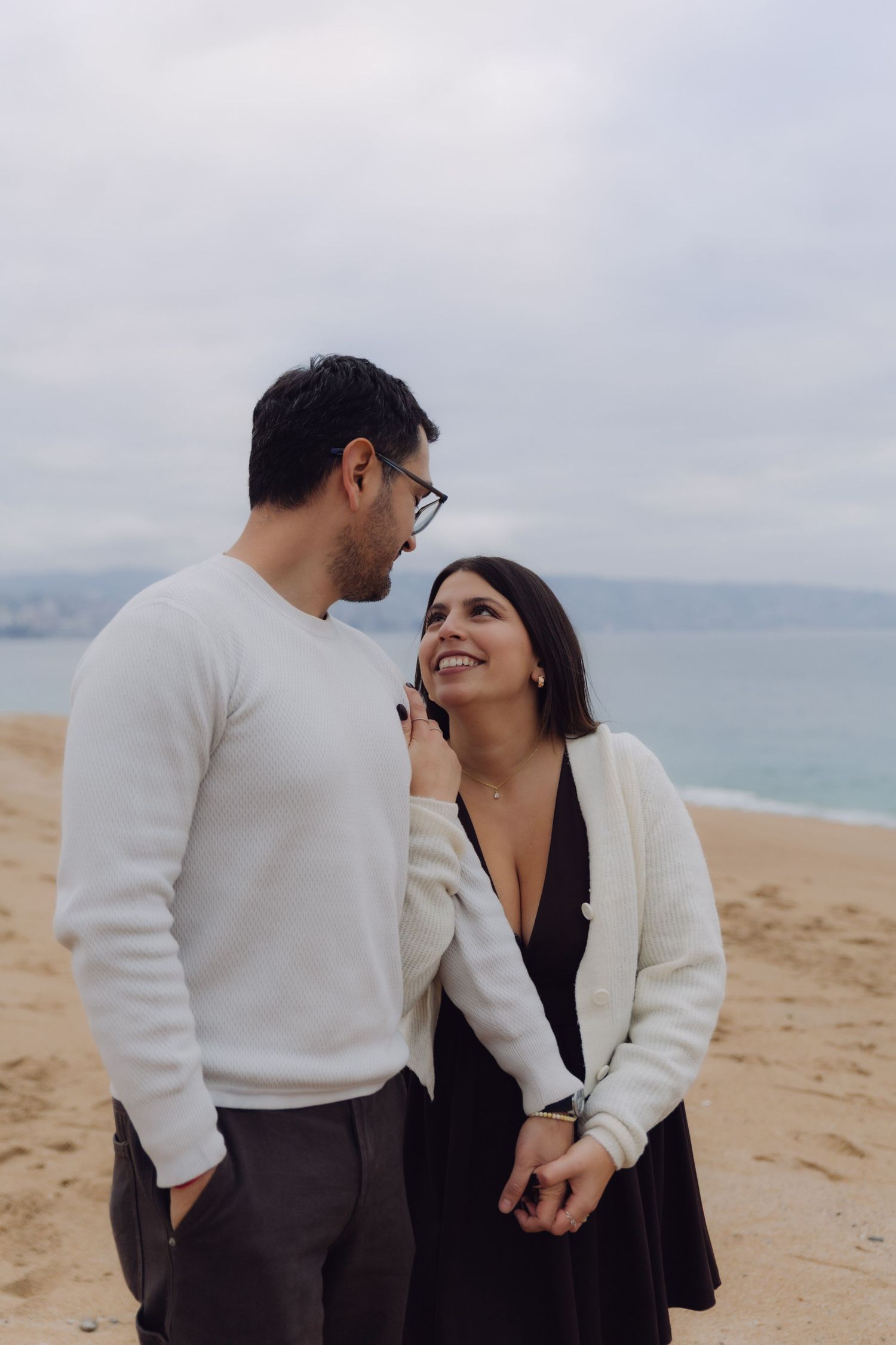 Smiling couple holding hands on a cloudy beach, woman in white cardigan looking up at man with glasses.