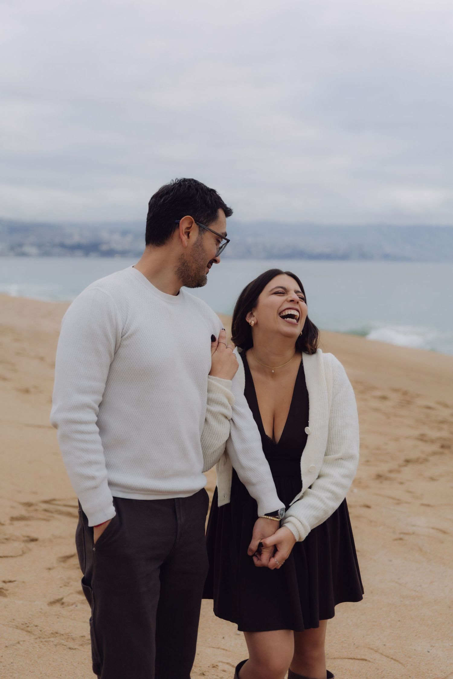 Laughing couple walking hand in hand on a sandy beach, woman in black dress and white cardigan.