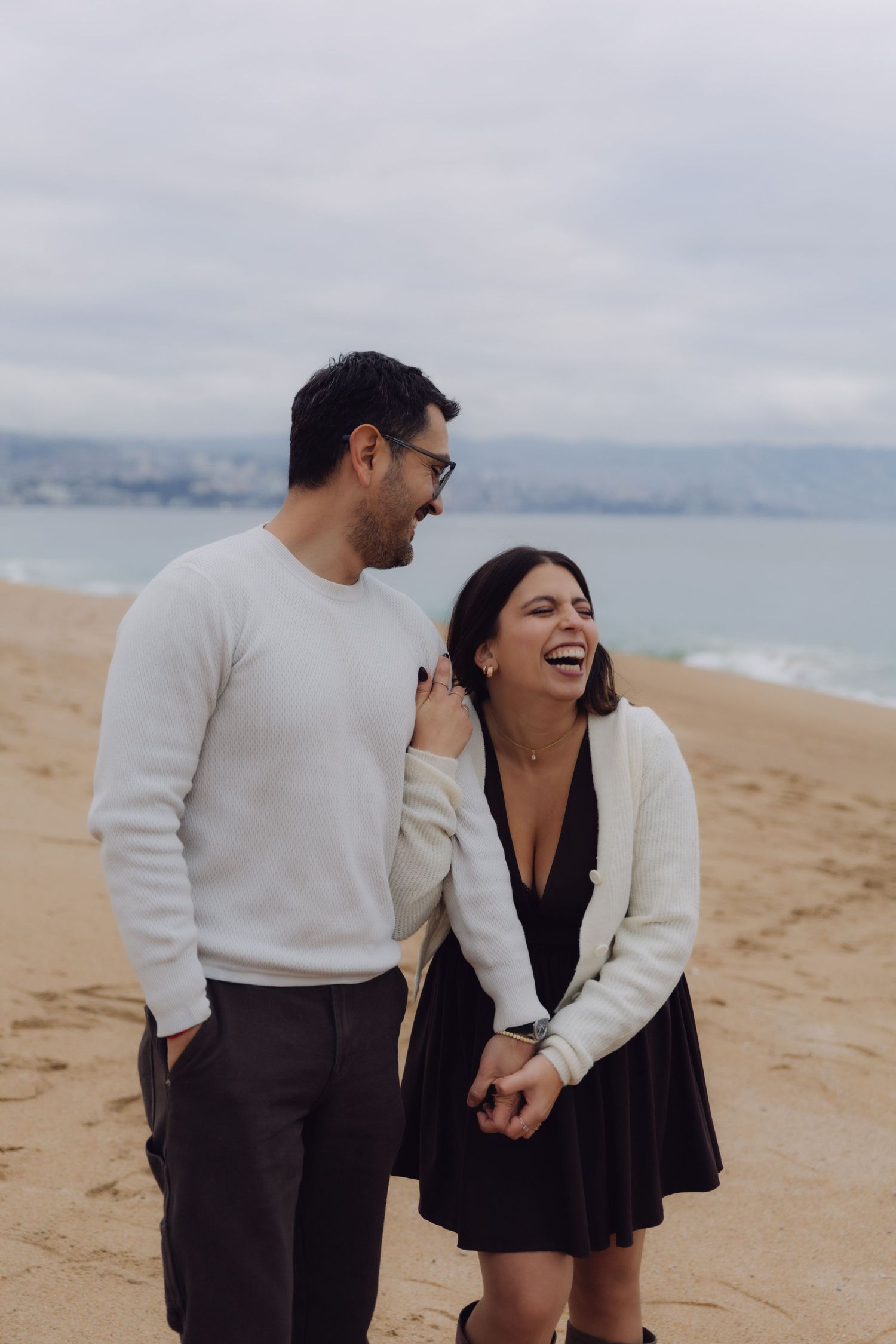 Couple sharing a joyful laugh while walking on a beach, woman in black dress holding mans hand.