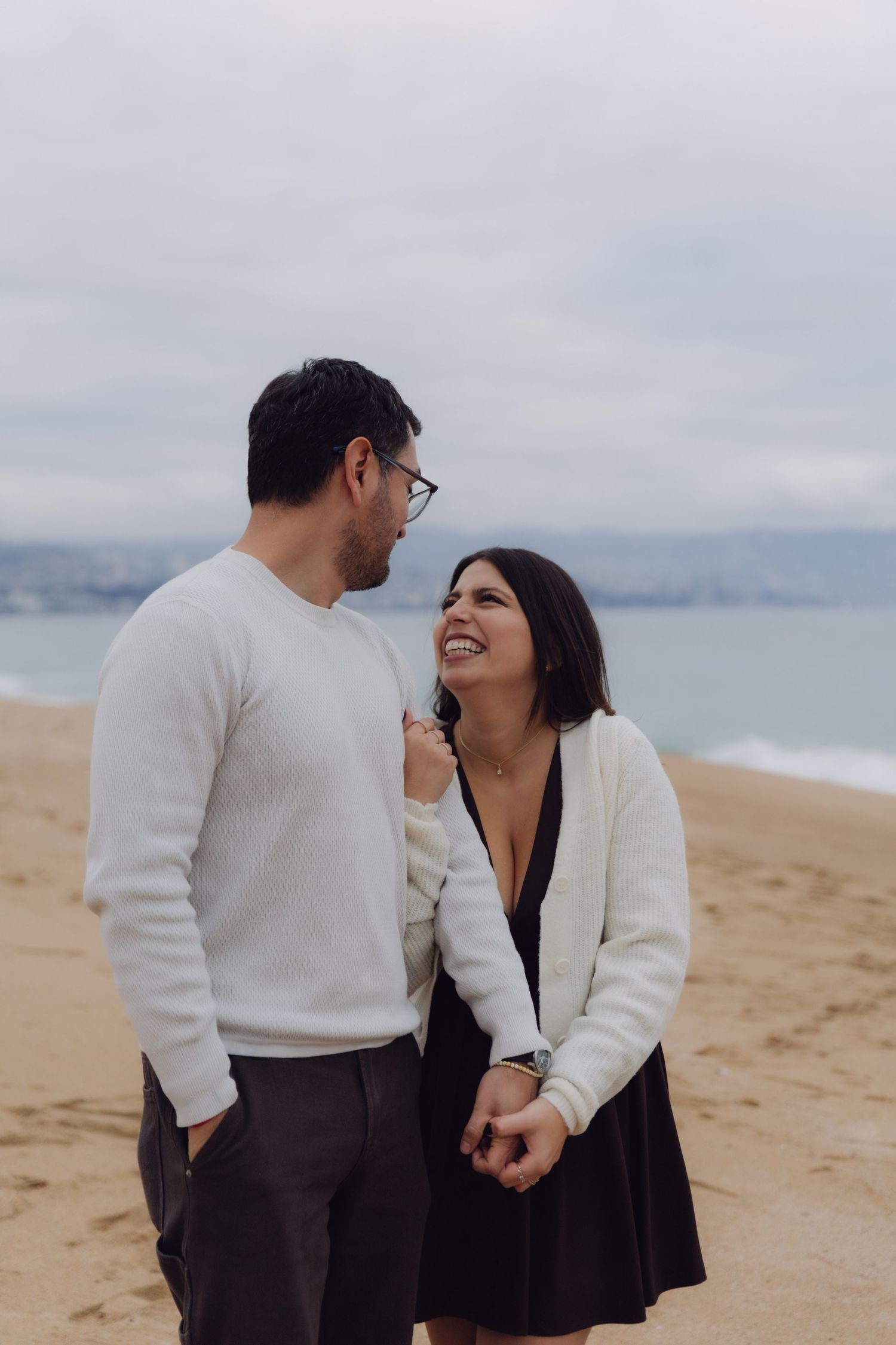 Couple smiling at each other on a sandy beach, woman in white cardigan and black dress holding hands.