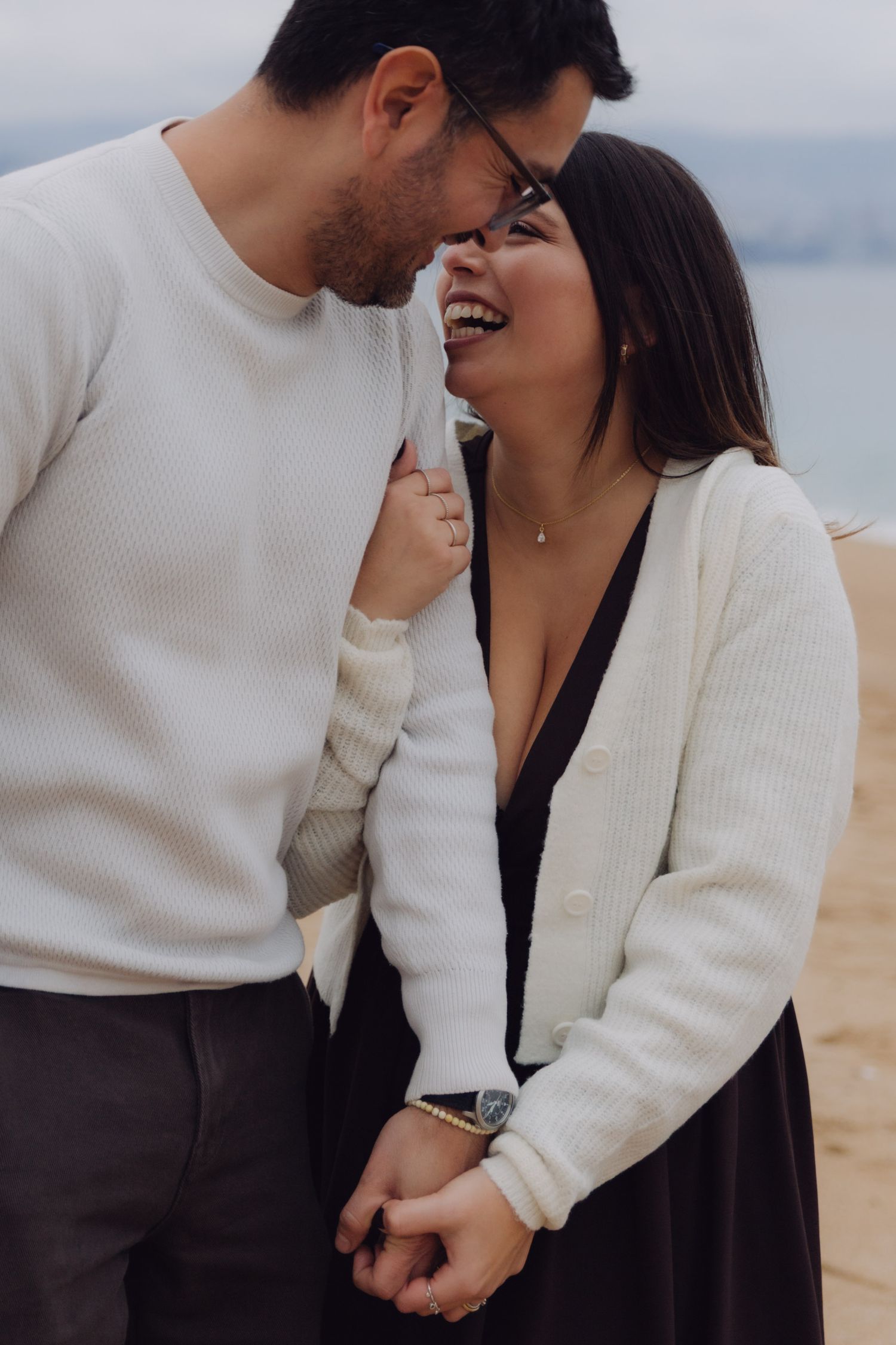 Close-up of laughing couple holding hands on a beach, woman in white cardigan and black dress.