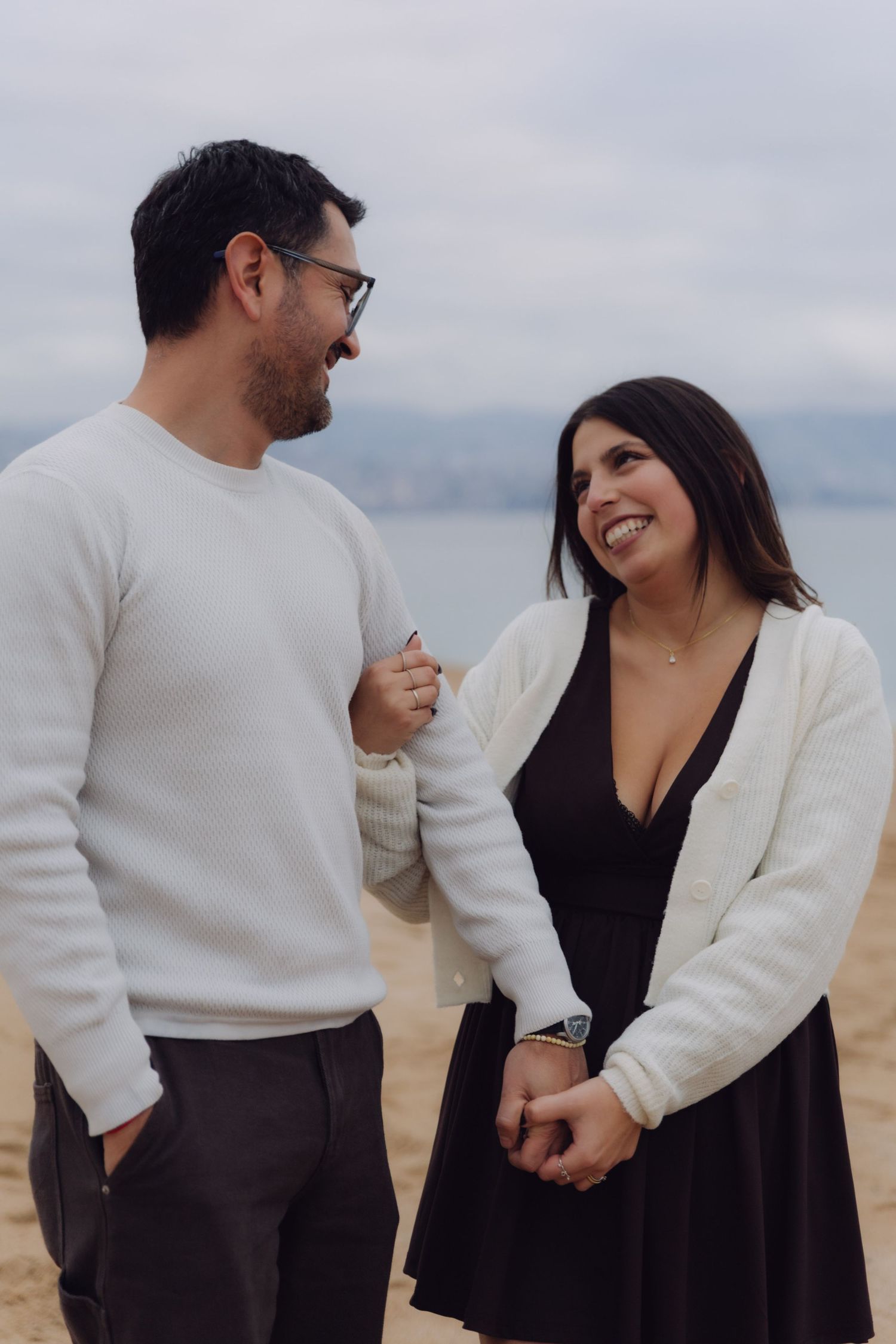 Happy couple smiling at each other on a sandy beach, woman in white cardigan and black dress.