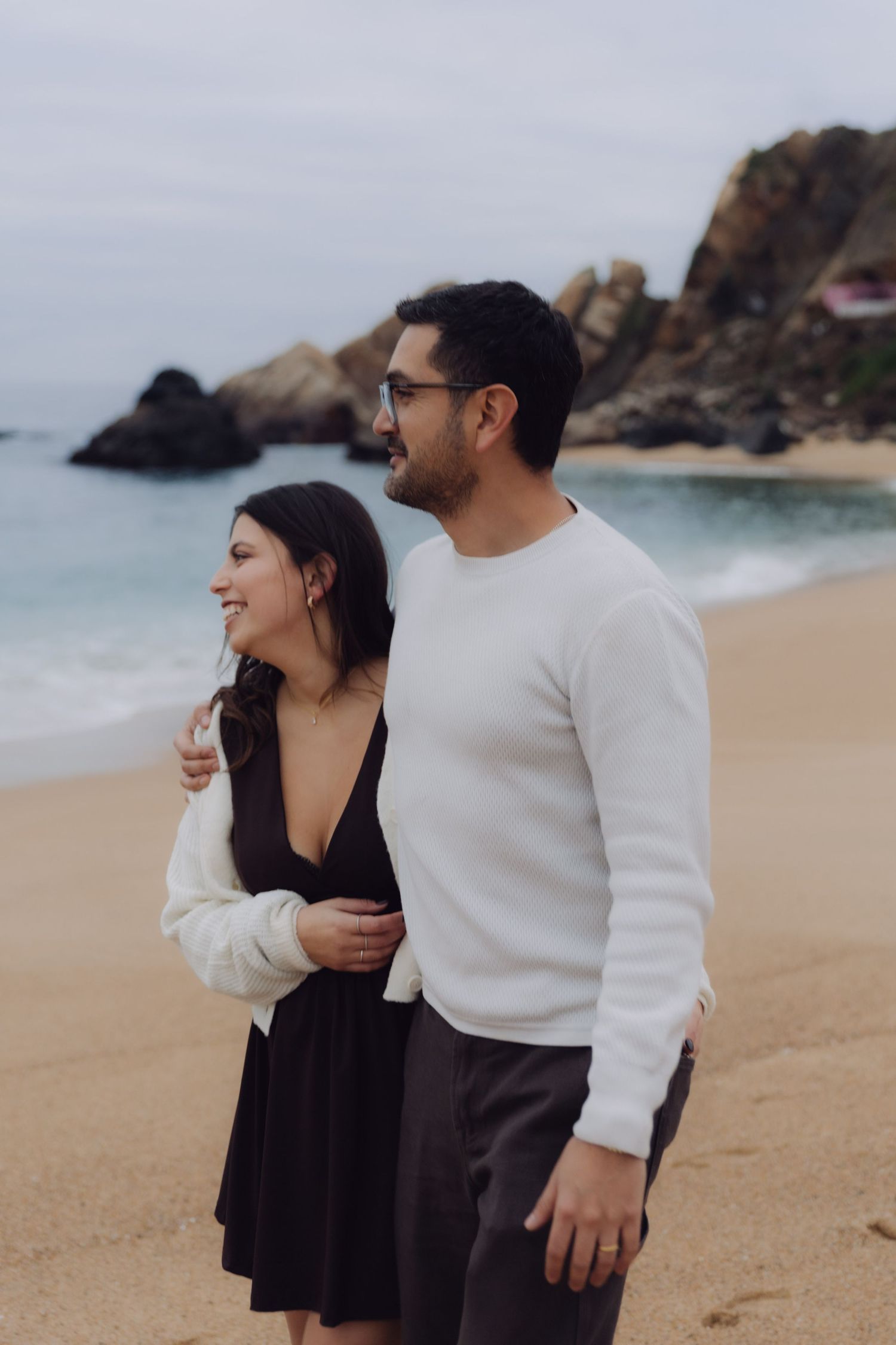 Couple embracing on a scenic beach with rocky cliffs, man in white sweater hugging woman in black dress.