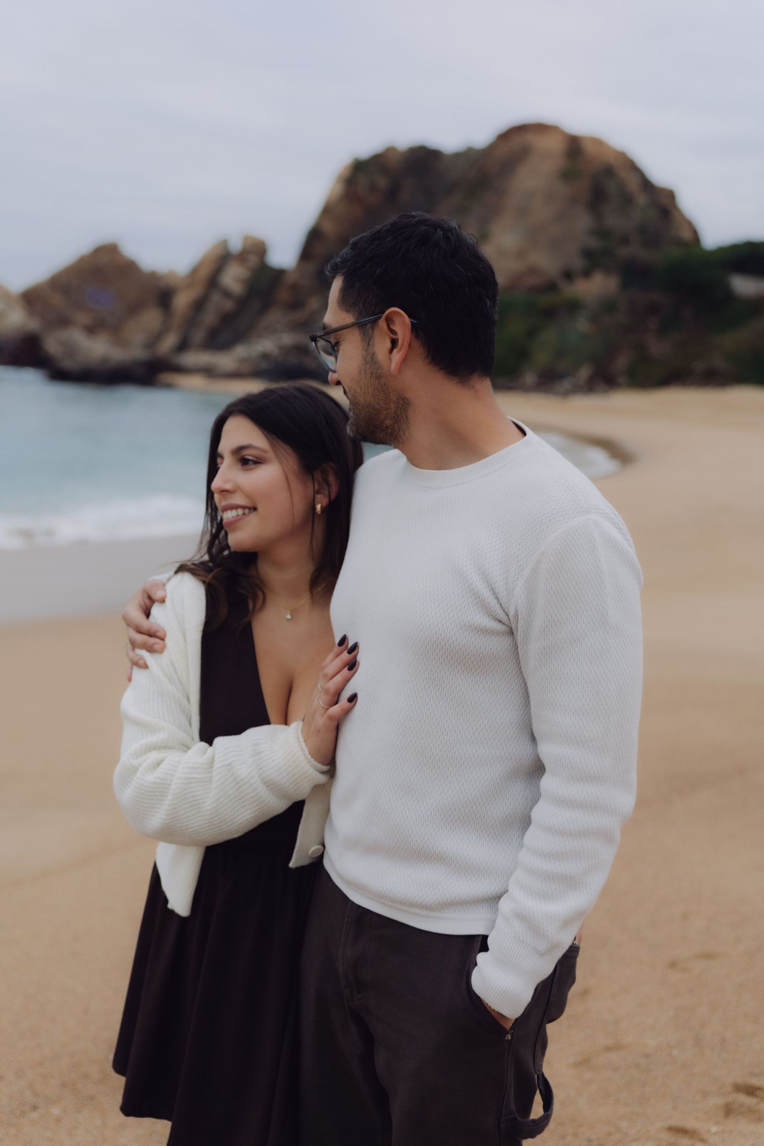 Couple standing together on a sandy beach with rocky backdrop, woman smiling in white cardigan and black dress.