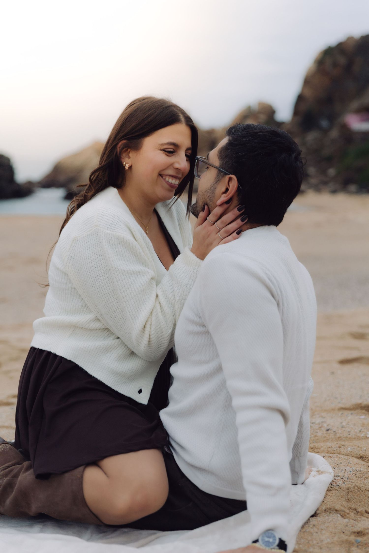 Couple sharing a romantic moment on sandy beach, woman laughing while holding mans face, both wearing white sweaters.