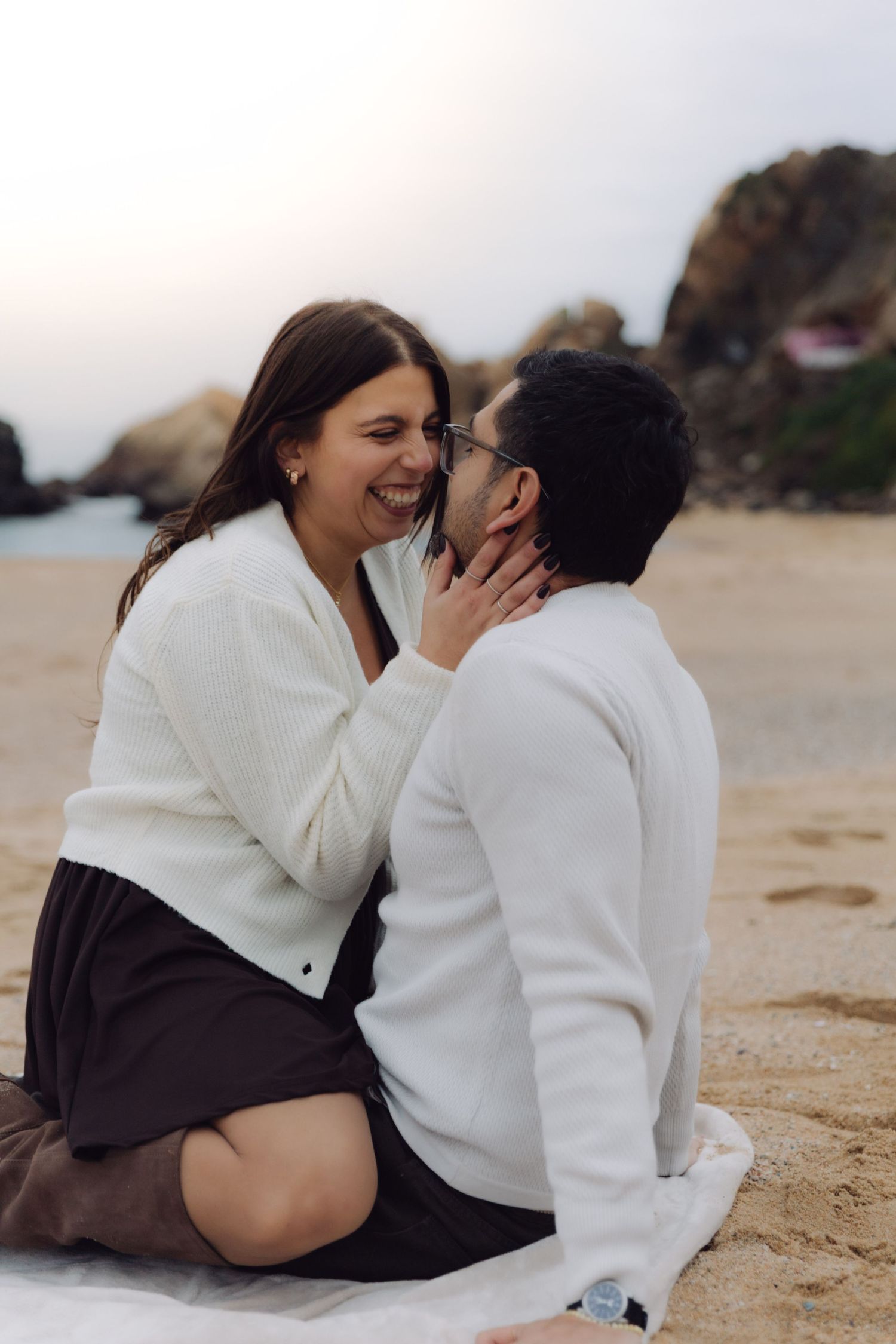 Happy couple sitting on beach blanket laughing together, woman touching mans face, wearing coordinating white outfits.