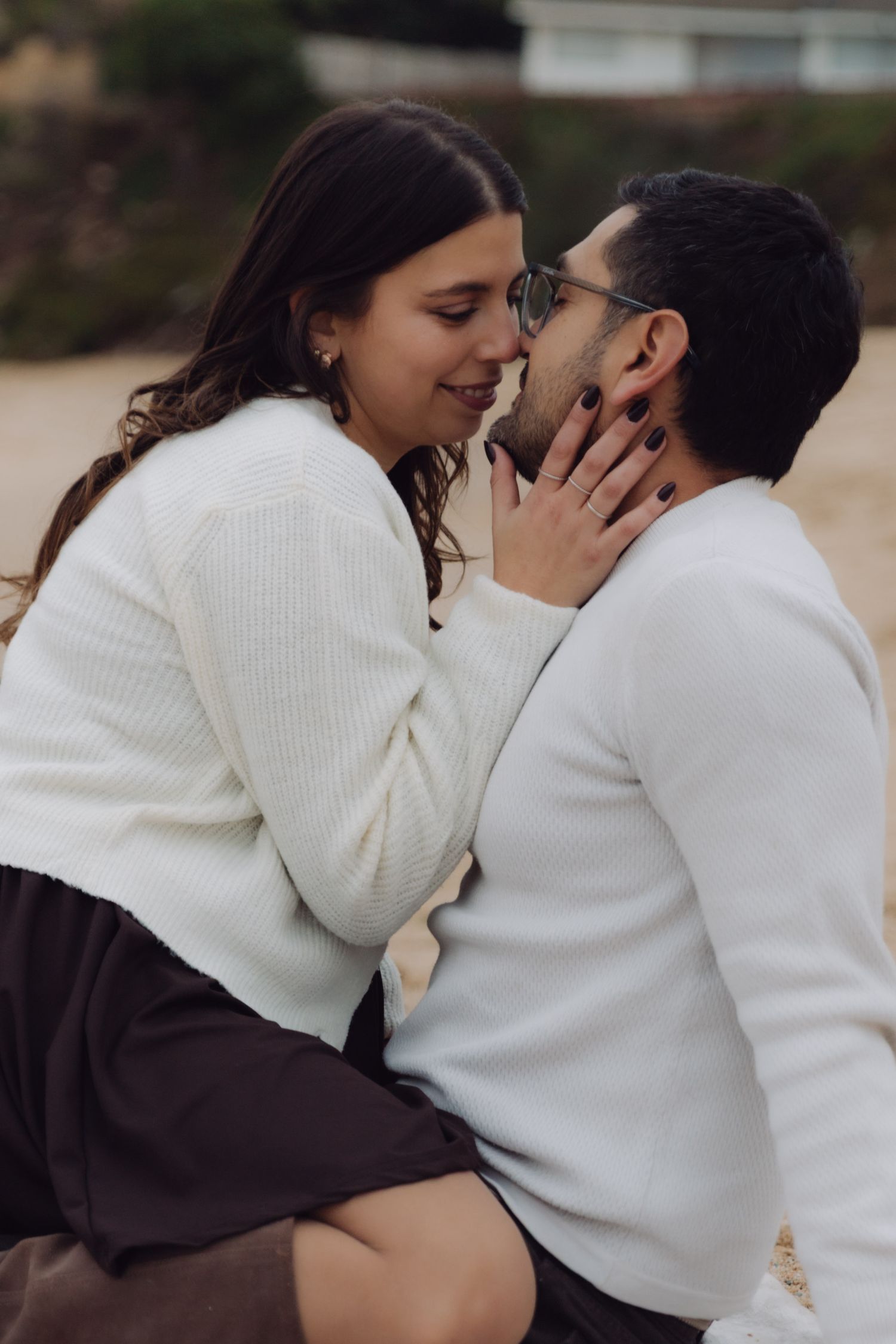 Romantic couple almost kissing on beach, woman in white cardigan and dark dress gently holding mans face close.