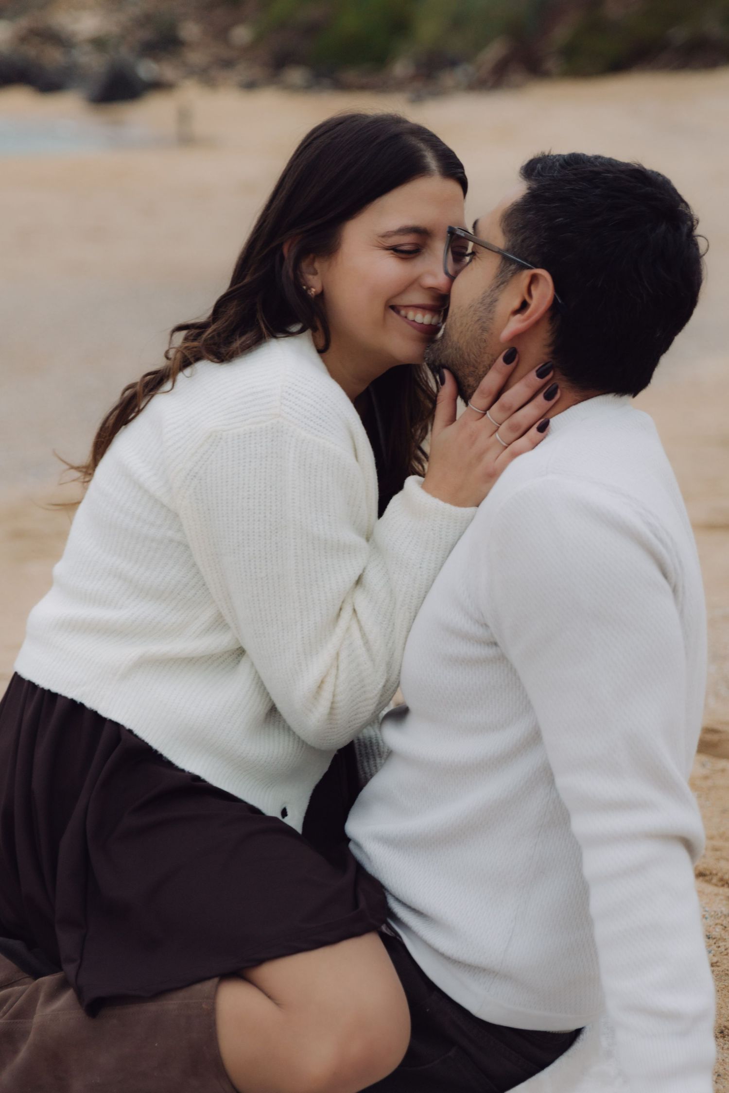 Smiling couple sharing a kiss on sandy beach, woman in white cardigan embracing man in white sweater outdoors.
