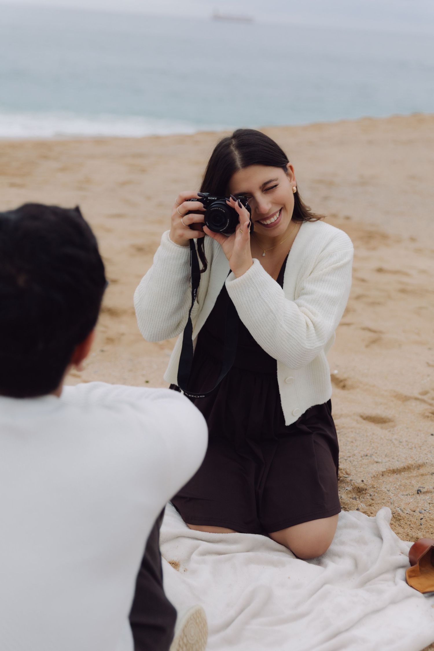 Smiling woman in white cardigan photographing man with film camera while sitting on blanket on sandy beach.