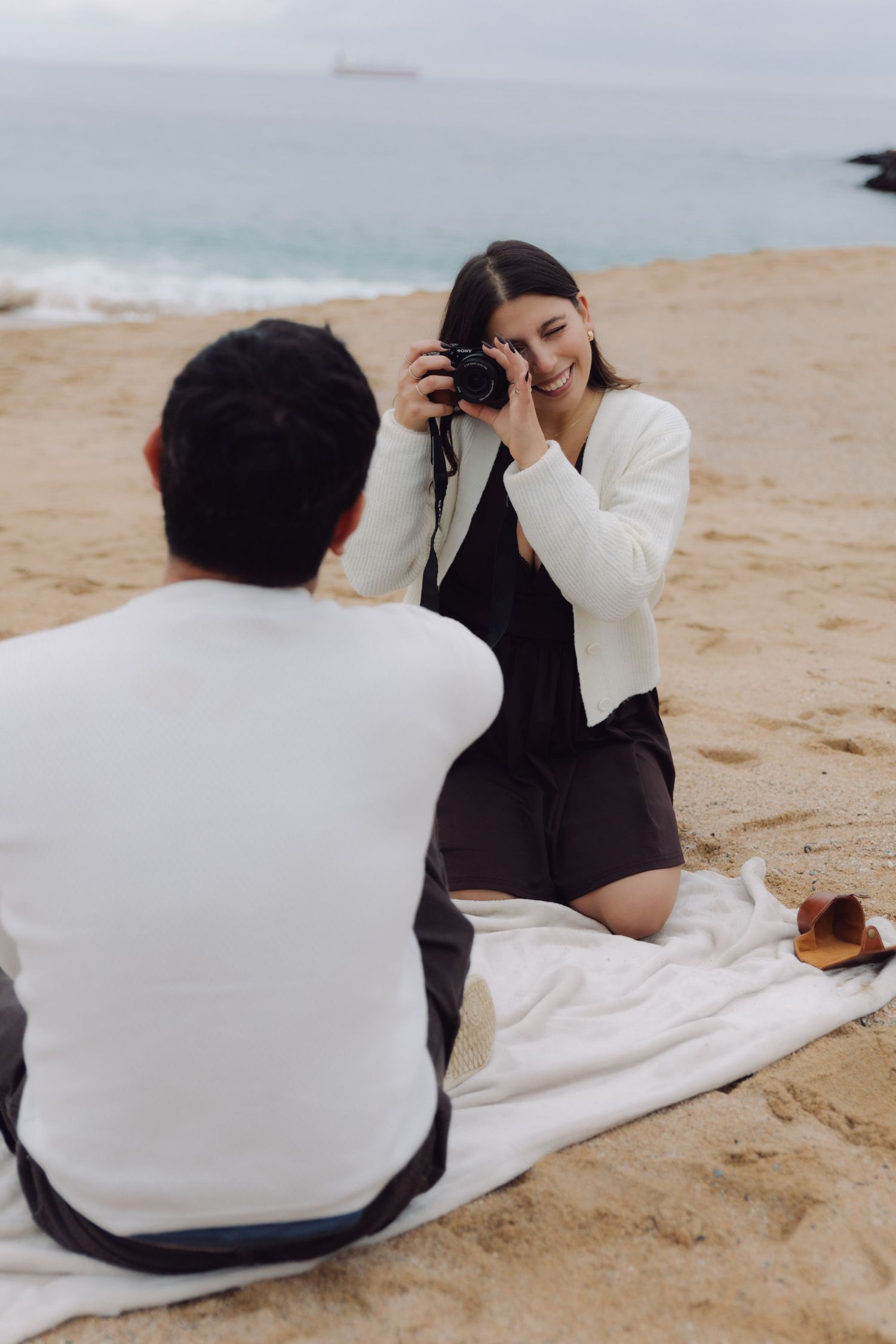 Young woman smiling while taking photo with camera of man sitting on white blanket on overcast beach.