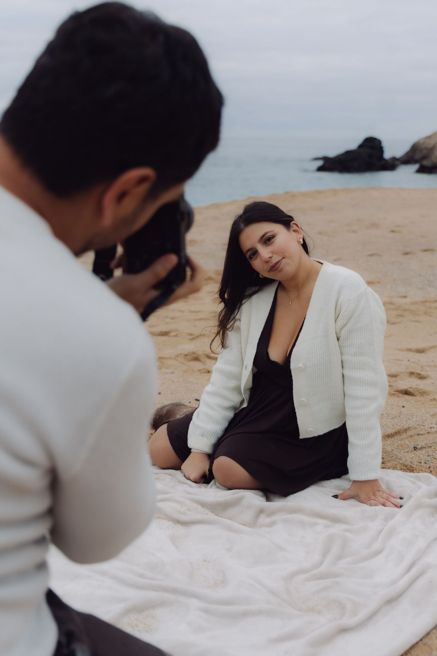 Man photographing woman in white cardigan and black dress sitting gracefully on blanket on sandy beach.