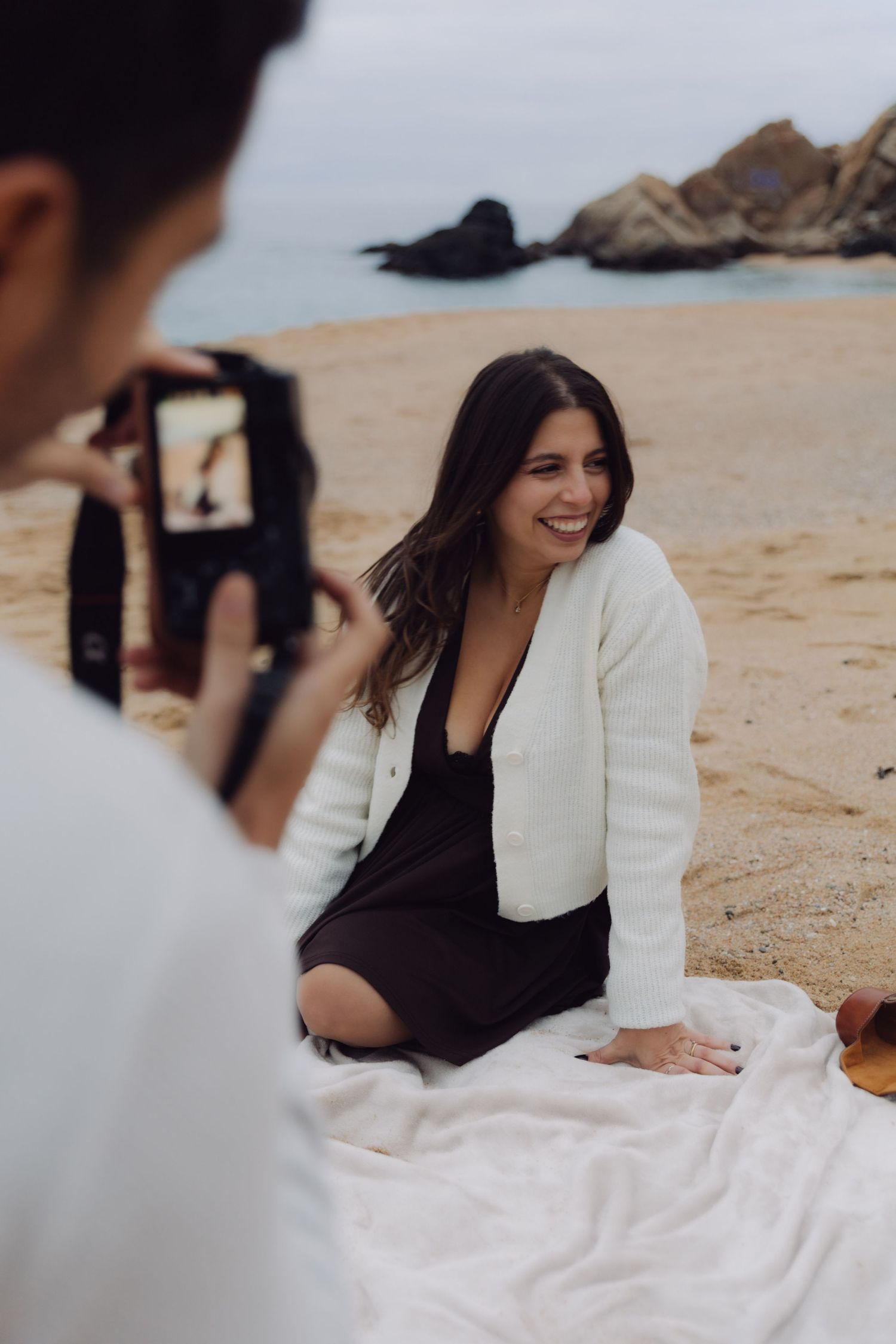 Man capturing photo of smiling woman in white cardigan sitting on beach blanket with rocky coastline behind her.