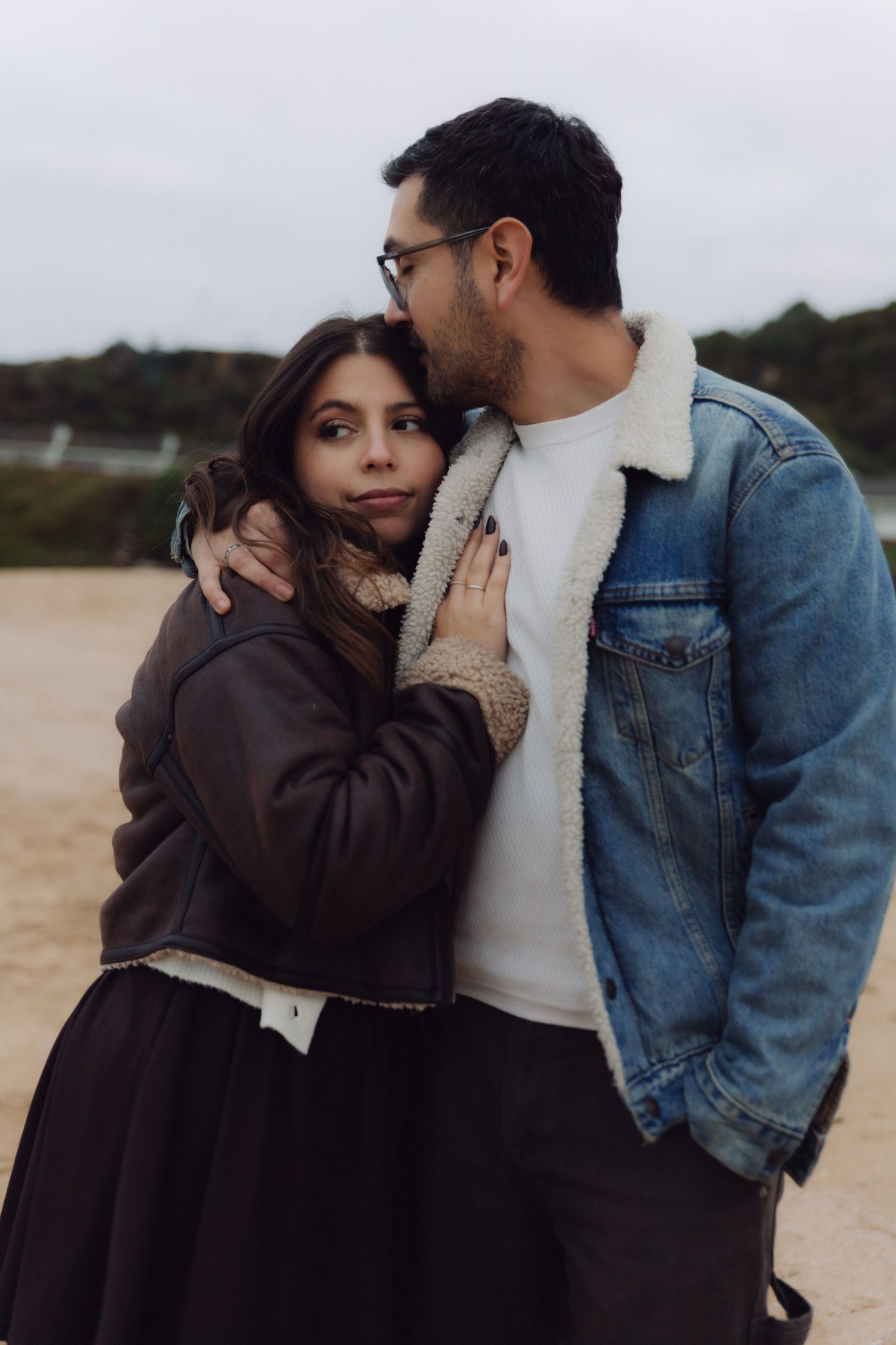 Couple embracing on sandy beach, man in denim sherpa jacket kissing womans head while she leans against him.