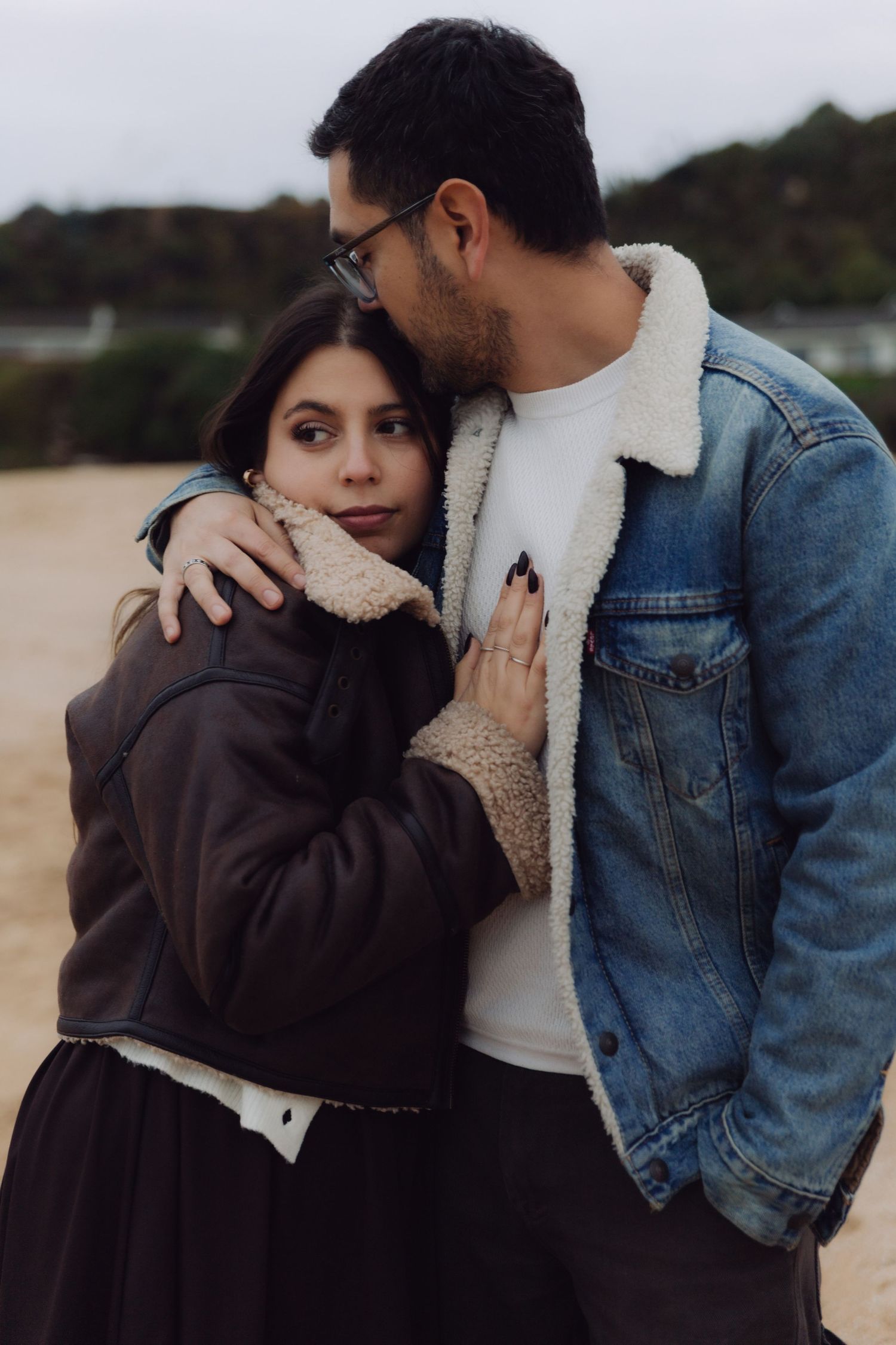 Romantic couple standing close on beach, woman in brown leather jacket leaning into man wearing denim sherpa jacket.