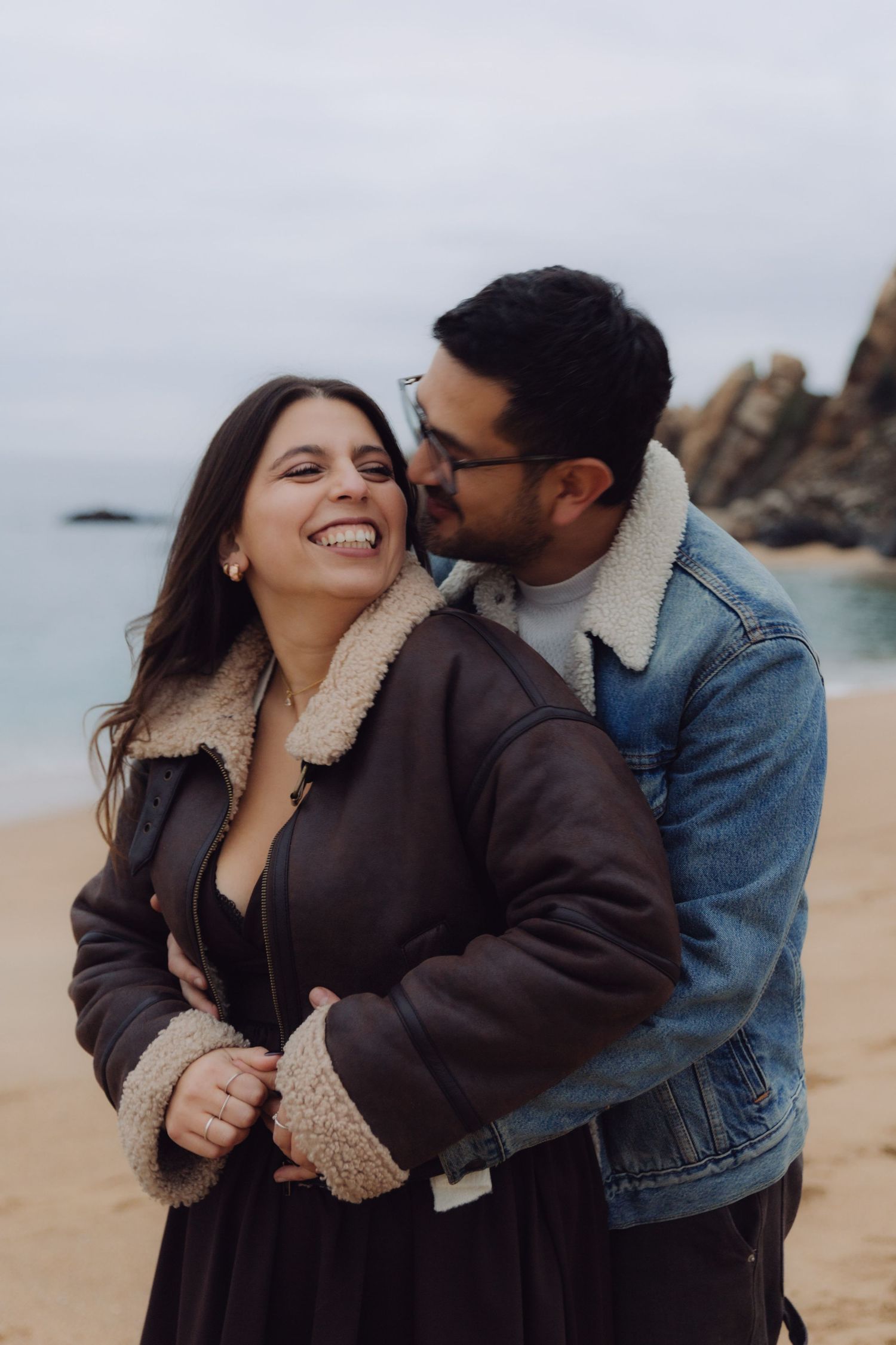 Couple embracing on a beach, woman laughing as man kisses her cheek, both wearing winter jackets.