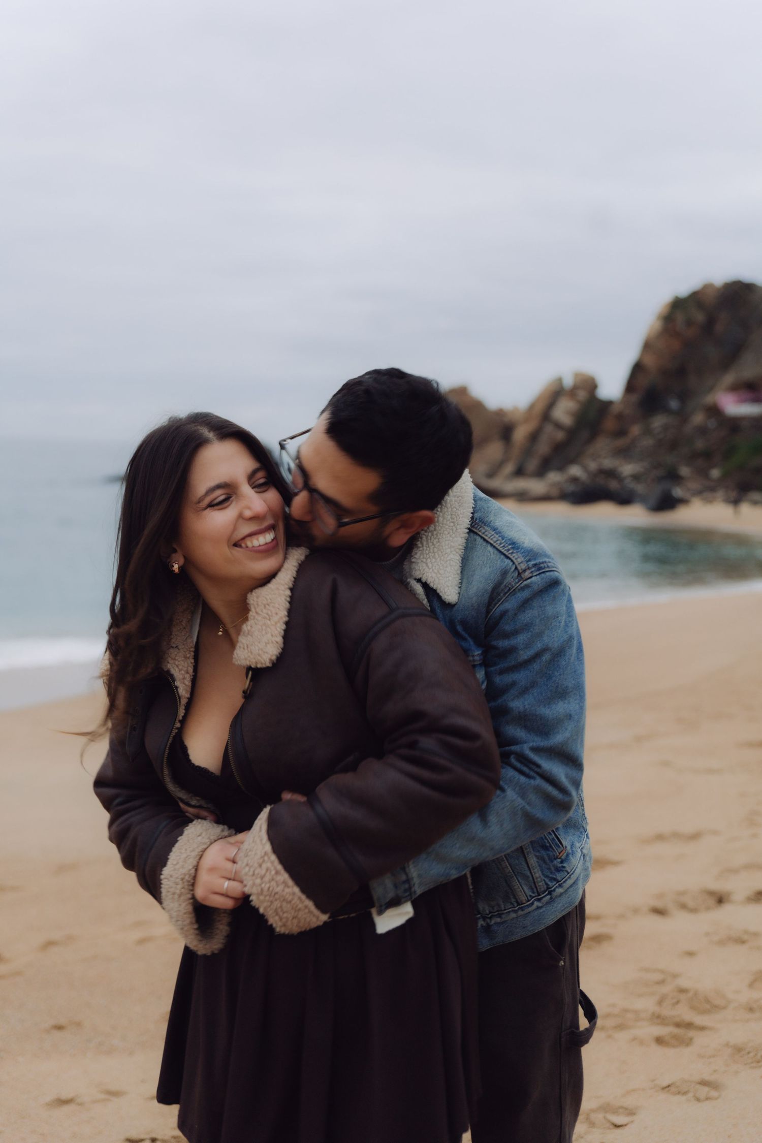 Happy couple hugging on a sandy beach, woman smiling as man kisses her cheek in cool weather.