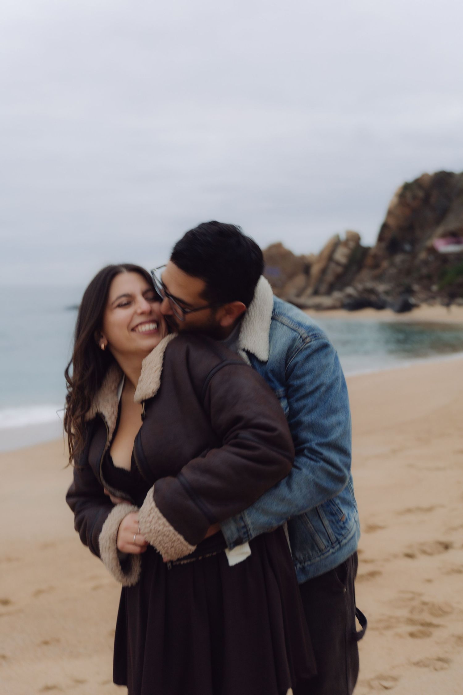 Romantic couple sharing a tender moment on a beach, woman smiling as man kisses her cheek.