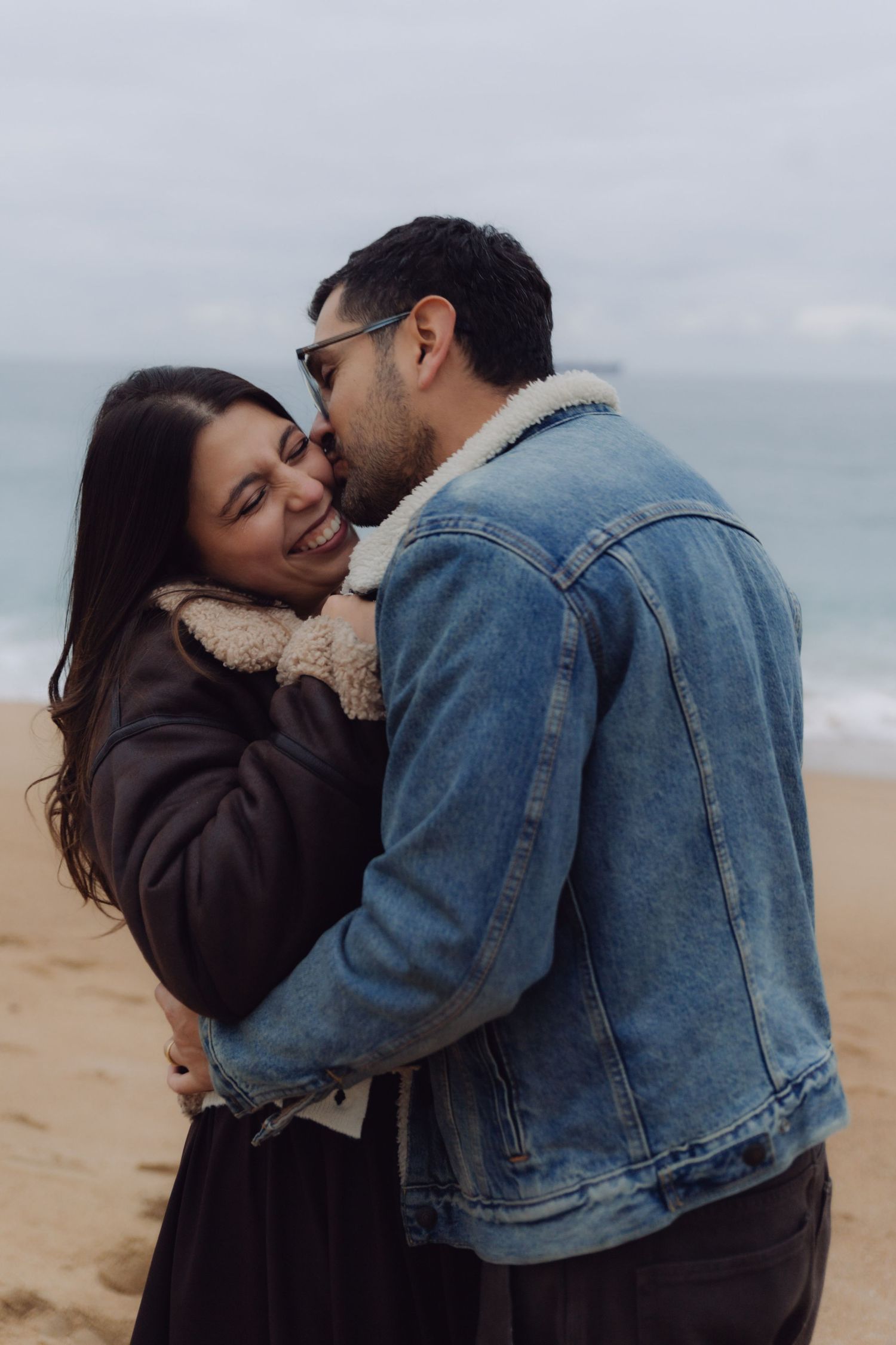 Smiling woman embracing man in denim jacket on a cloudy beach, sharing a romantic moment.