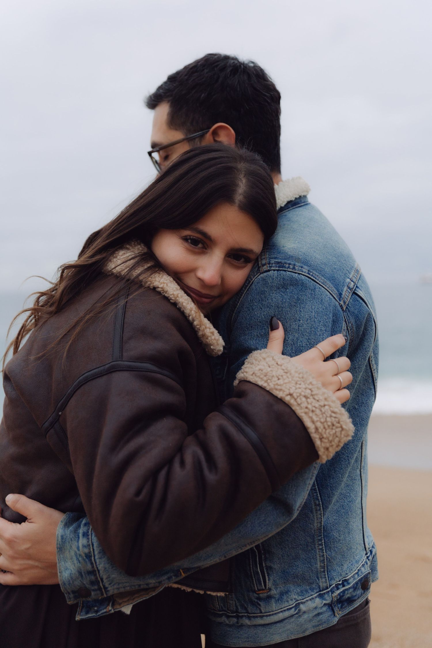 Couple sharing a warm hug on a beach, woman in brown shearling coat embracing man in denim jacket.