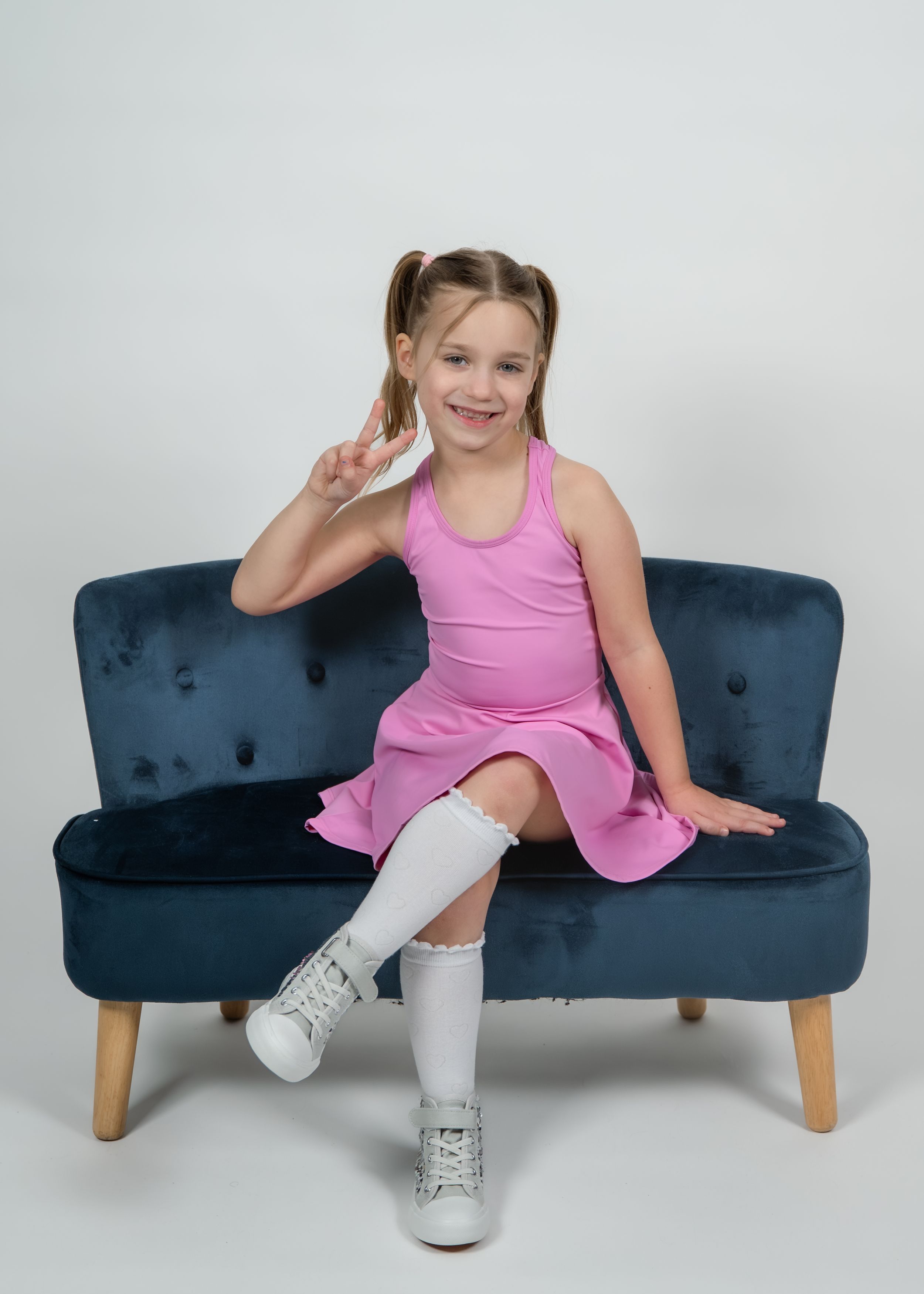 Young girl in pink dress and white knee socks sits on blue velvet sofa, smiling and making a peace sign.