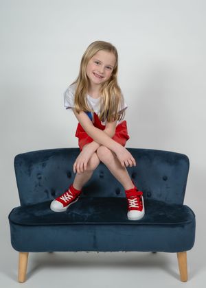 Young blonde girl sitting on a navy velvet sofa, wearing white tee, red skirt, and red sneakers, smiling at camera.