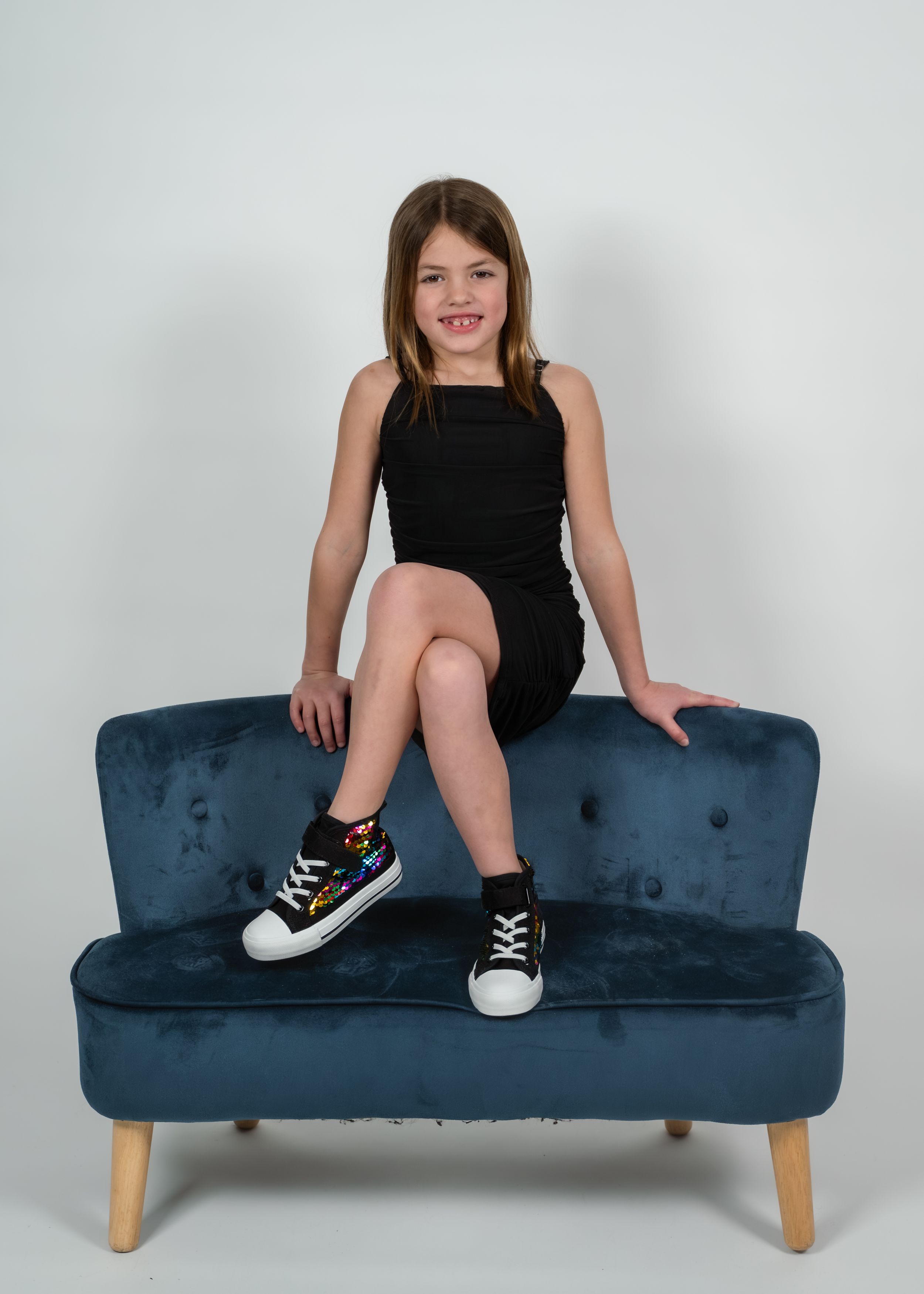 Young girl in black dress and high-top sneakers sitting on a teal velvet mini sofa against white background.
