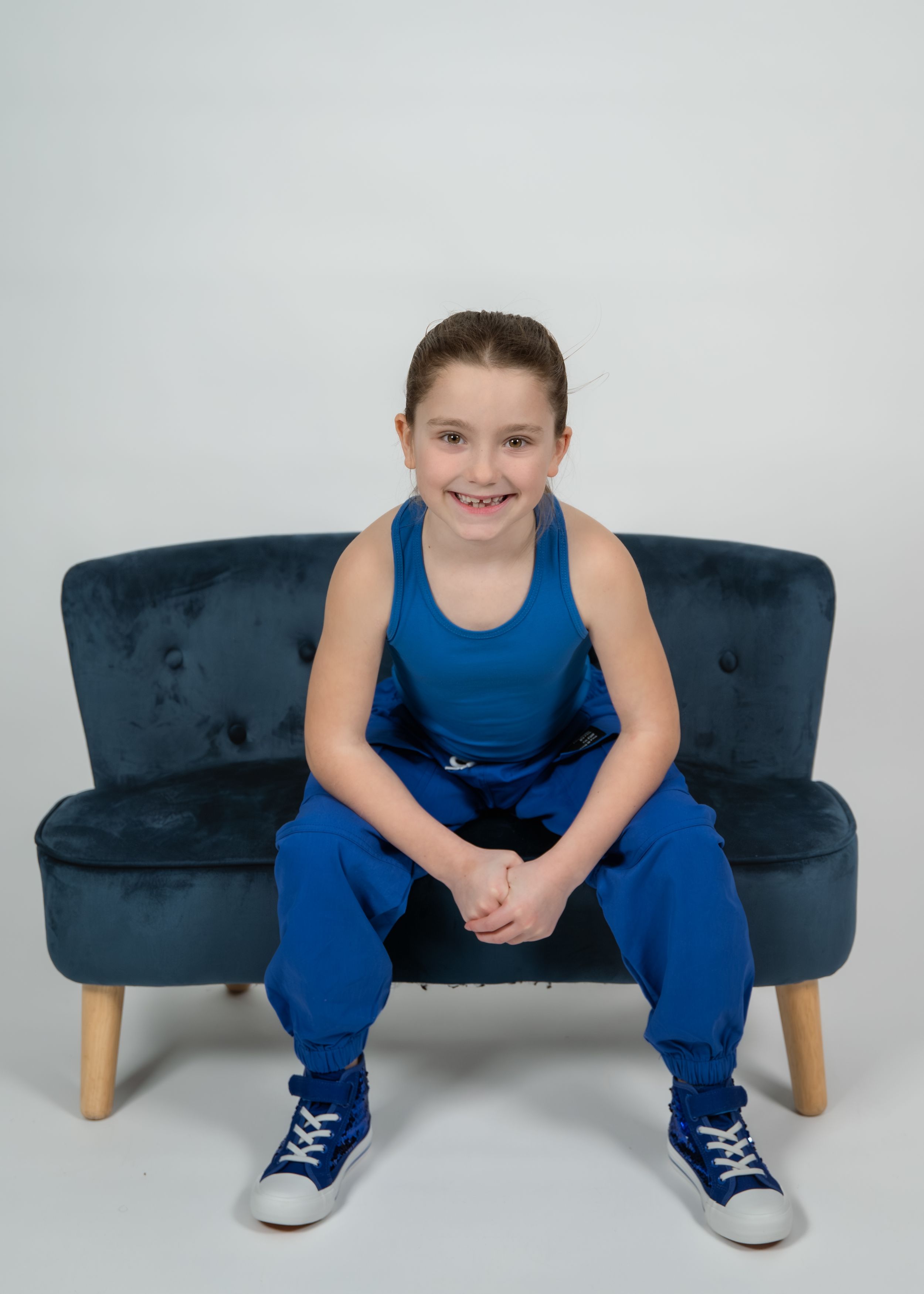 A smiling child in a blue outfit sits on a small navy velvet sofa against a white background.