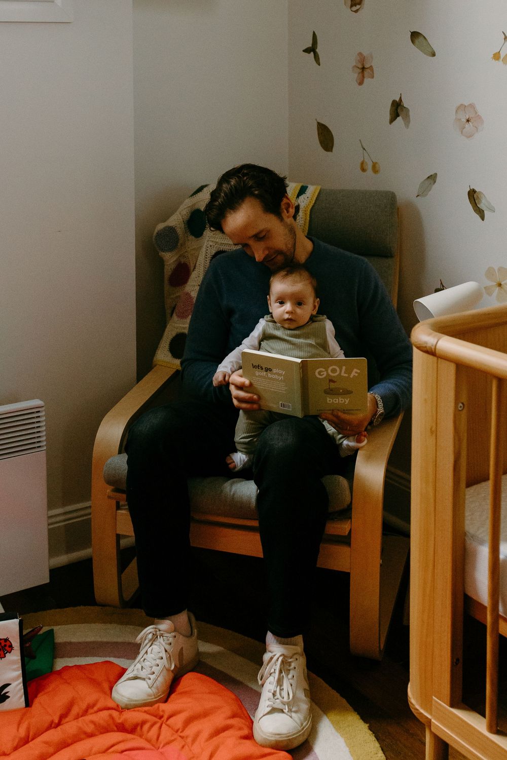 Father reading a golf book to his newborn baby while sitting in a nursery chair beside a wooden crib.