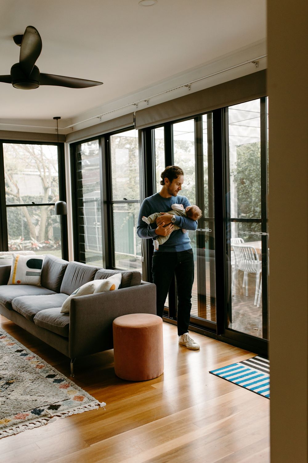 Young father in blue sweater cradling newborn baby while standing in modern living room by large windows.