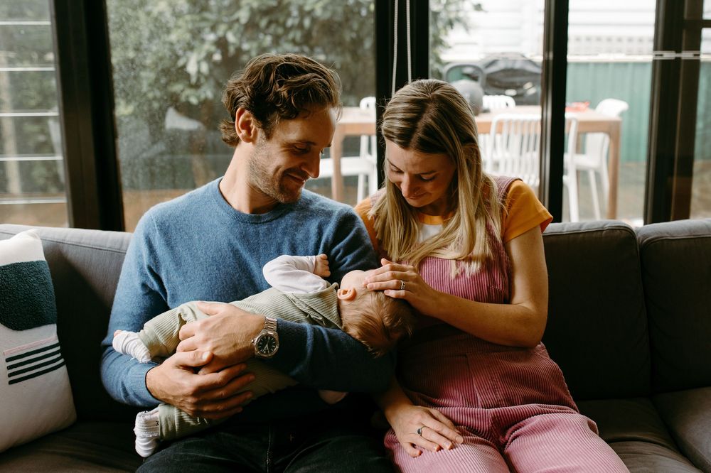 A couple sitting on a gray couch lovingly gazing at their newborn baby in a cozy home setting.